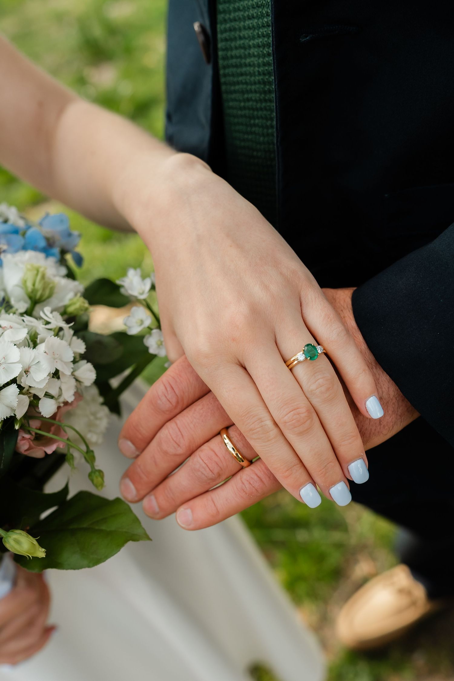 Close-up of newlyweds hands showing gold wedding bands and emerald engagement ring with bouquet.