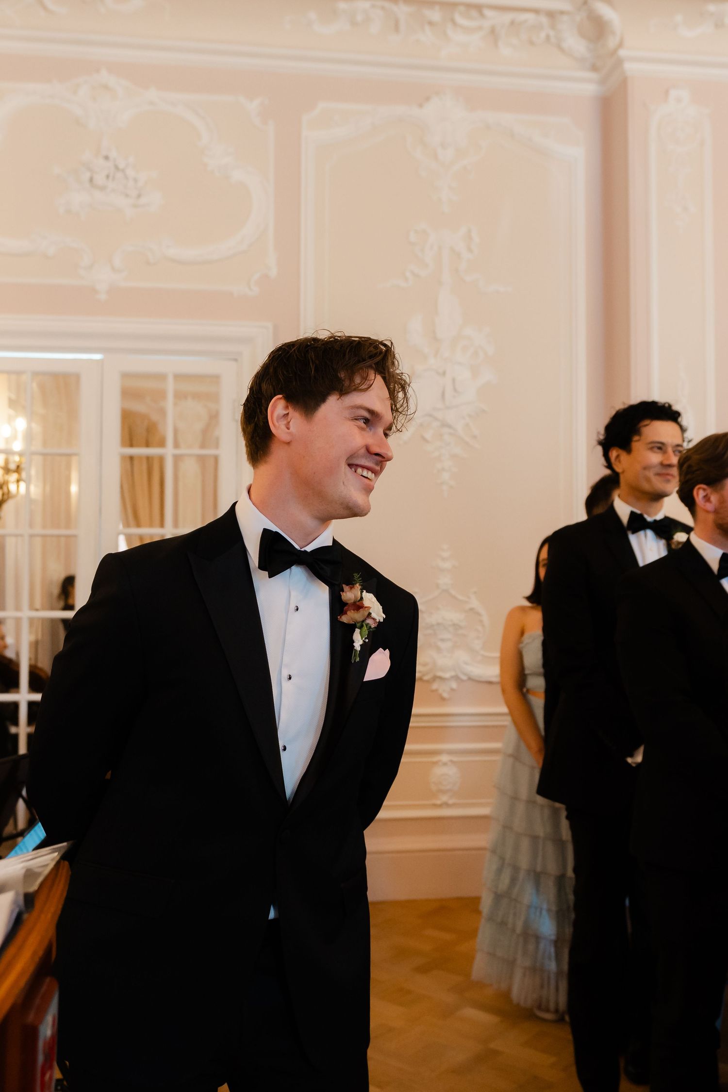 Handsome groom in black tuxedo with boutonniere smiling during wedding ceremony in ornate venue.
