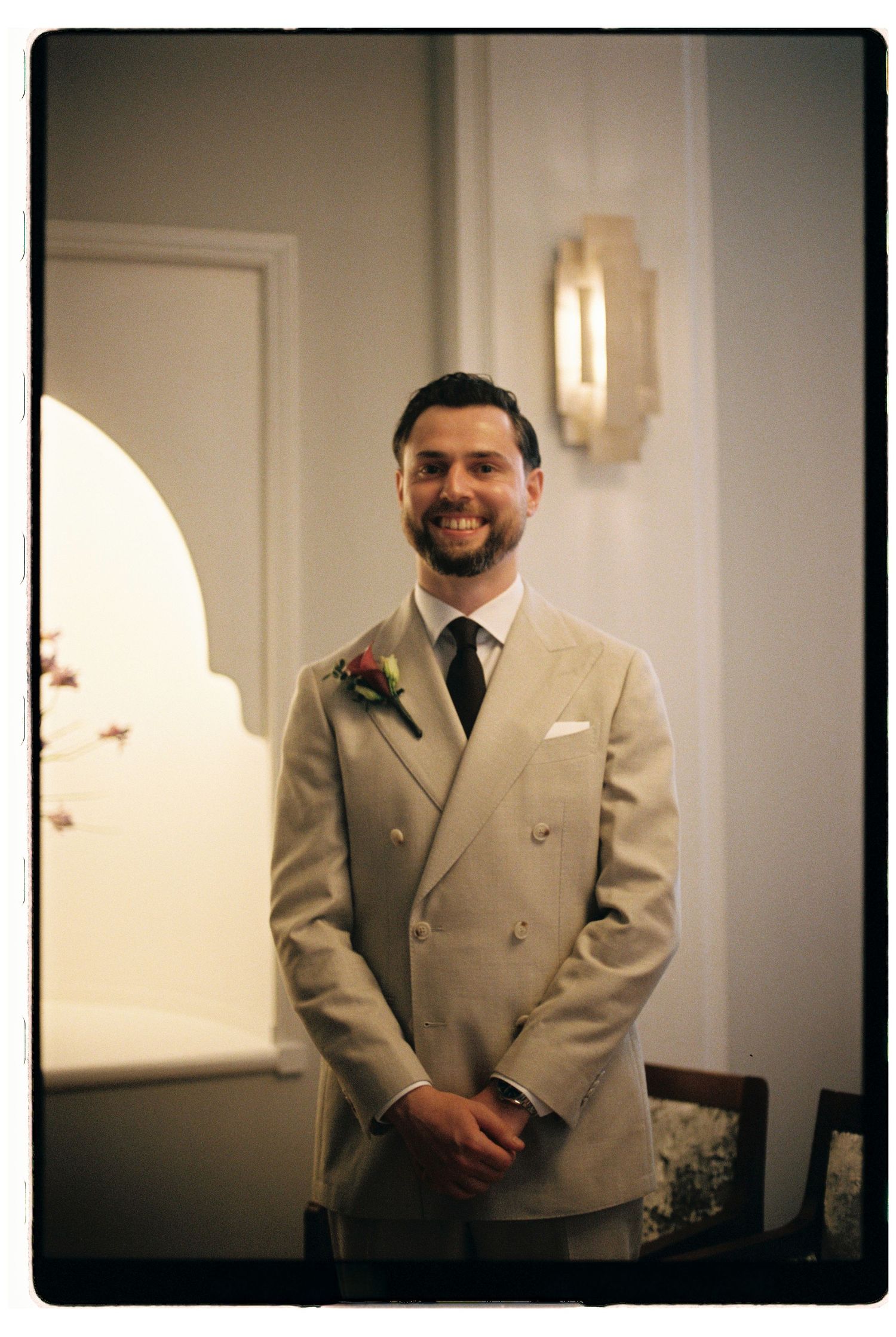 Groom in beige double-breasted suit with boutonniere smiling at wedding.