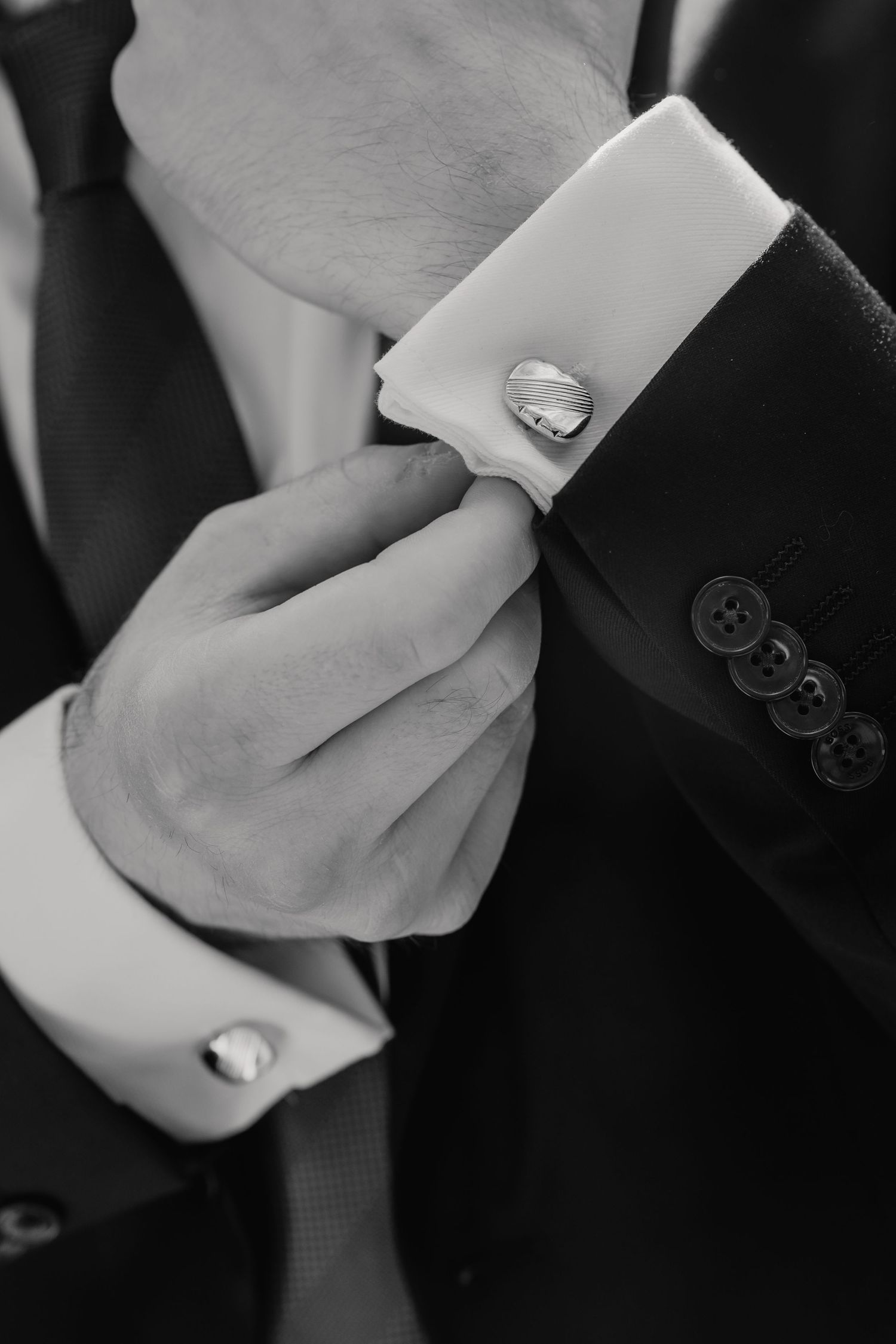 Groom adjusting silver cufflinks on white dress shirt sleeve in black and white wedding photo.