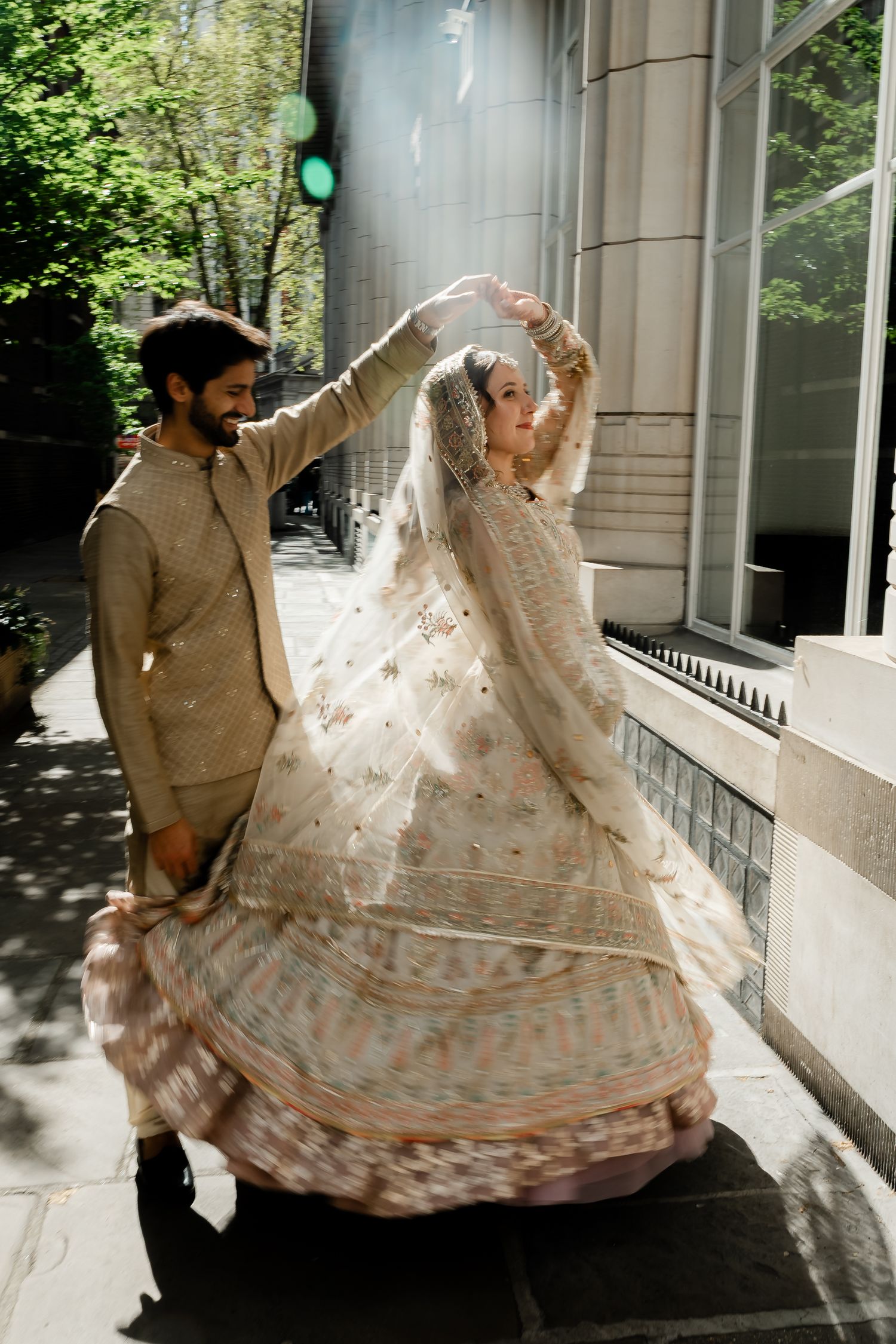 South Asian groom spinning bride in embroidered ivory lehenga on sunlit city sidewalk after wedding ceremony.
