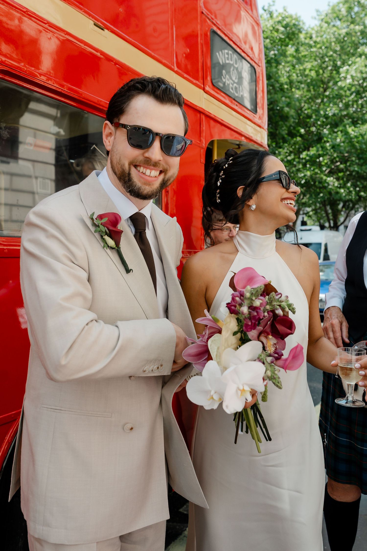 Stylish bride and groom in sunglasses smiling outside a red London double-decker bus, bride holding white and pink bouquet.