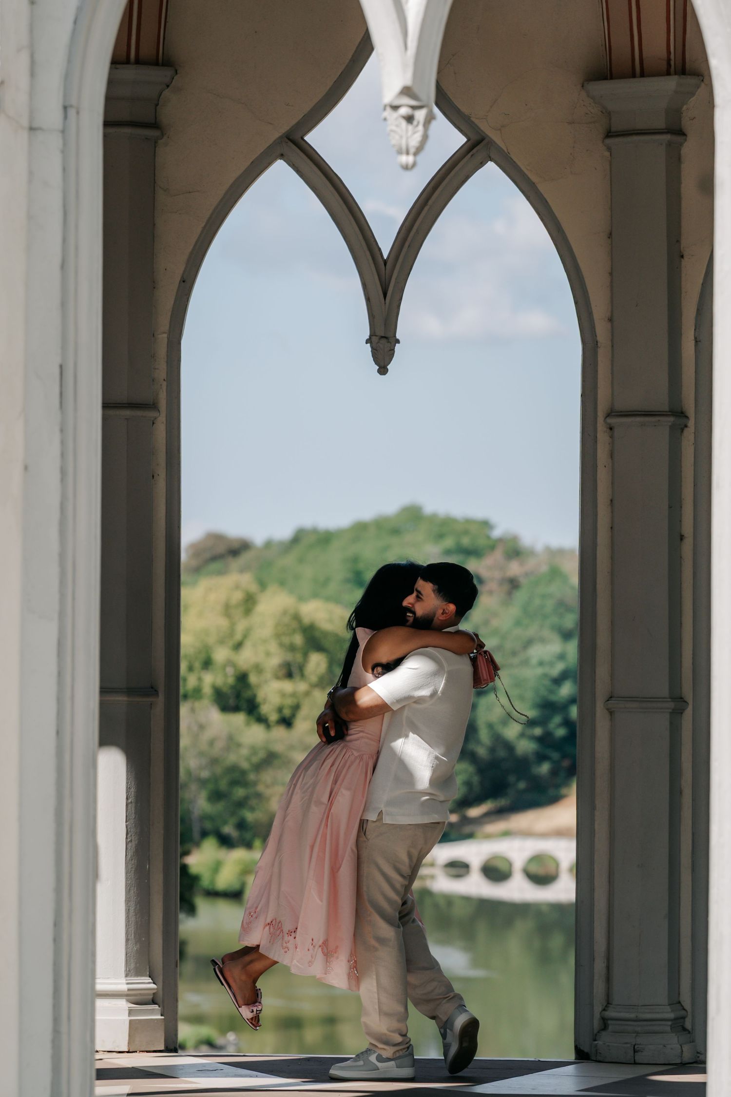 Couple embracing and spinning joyfully under elegant Gothic stone arch with scenic lake view.