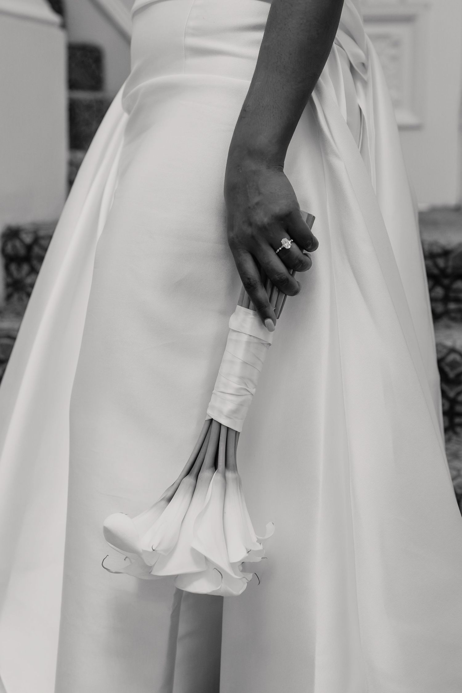Black and white photo of a bride in a white gown holding a calla lily bouquet with an engagement ring visible.