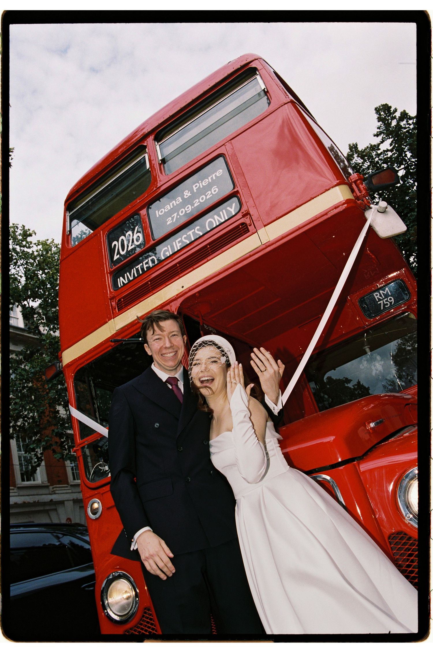 A joyful bride and groom pose in front of a classic red London double-decker wedding bus marked 2026.