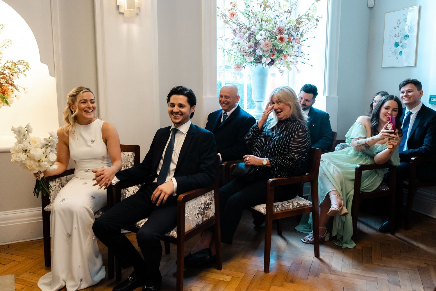 Bride and groom laugh together seated at wedding ceremony with smiling guests behind them.