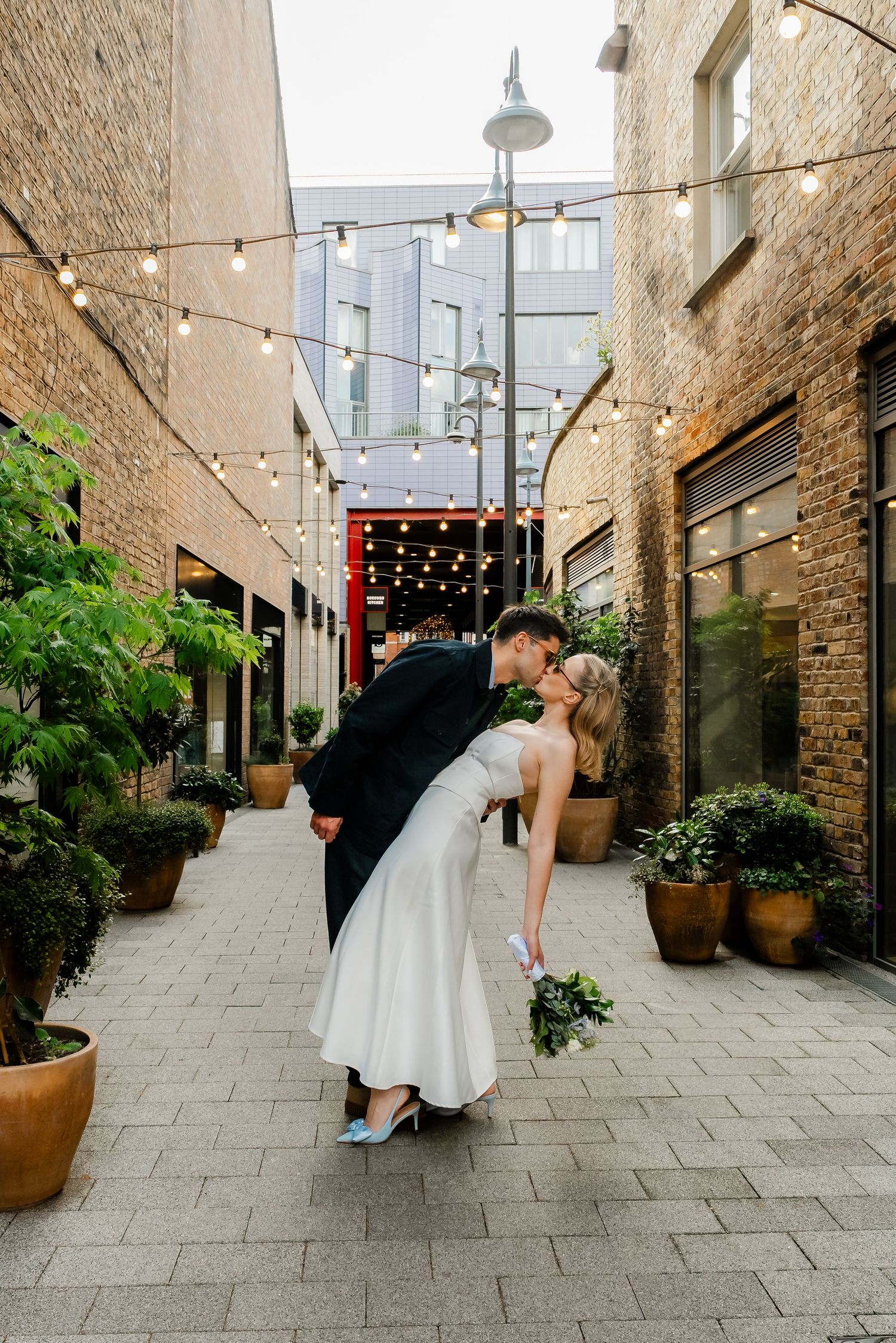 Groom dipping bride for a romantic kiss in a charming alley with string lights.