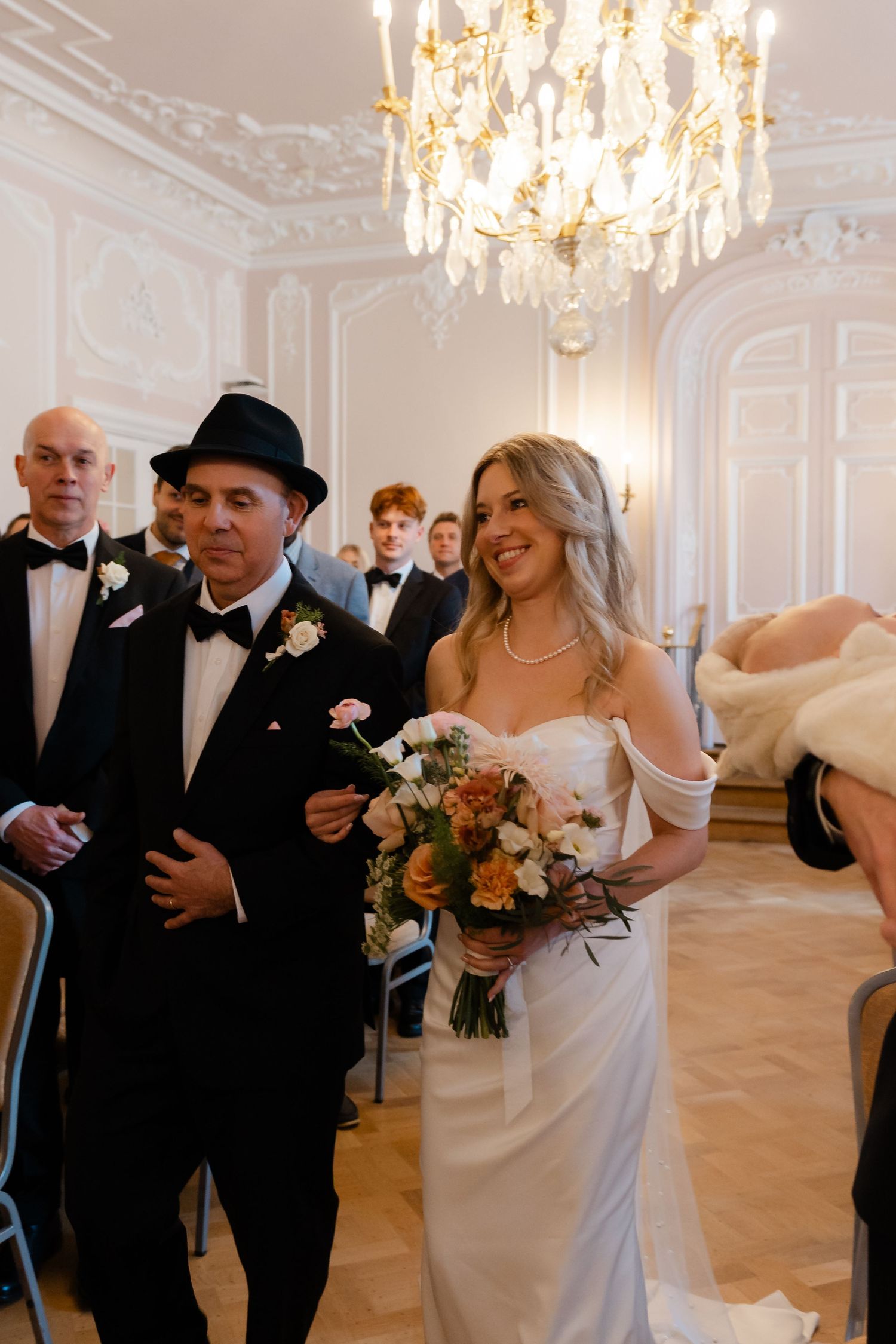 Smiling bride in white off-shoulder gown walks down the aisle with escort in black tuxedo.
