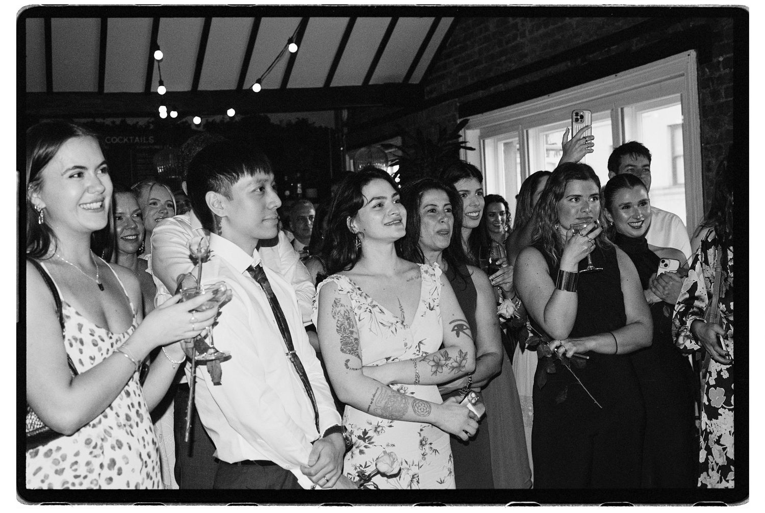 Black and white photo of wedding guests watching ceremony attentively holding drinks in decorated venue.