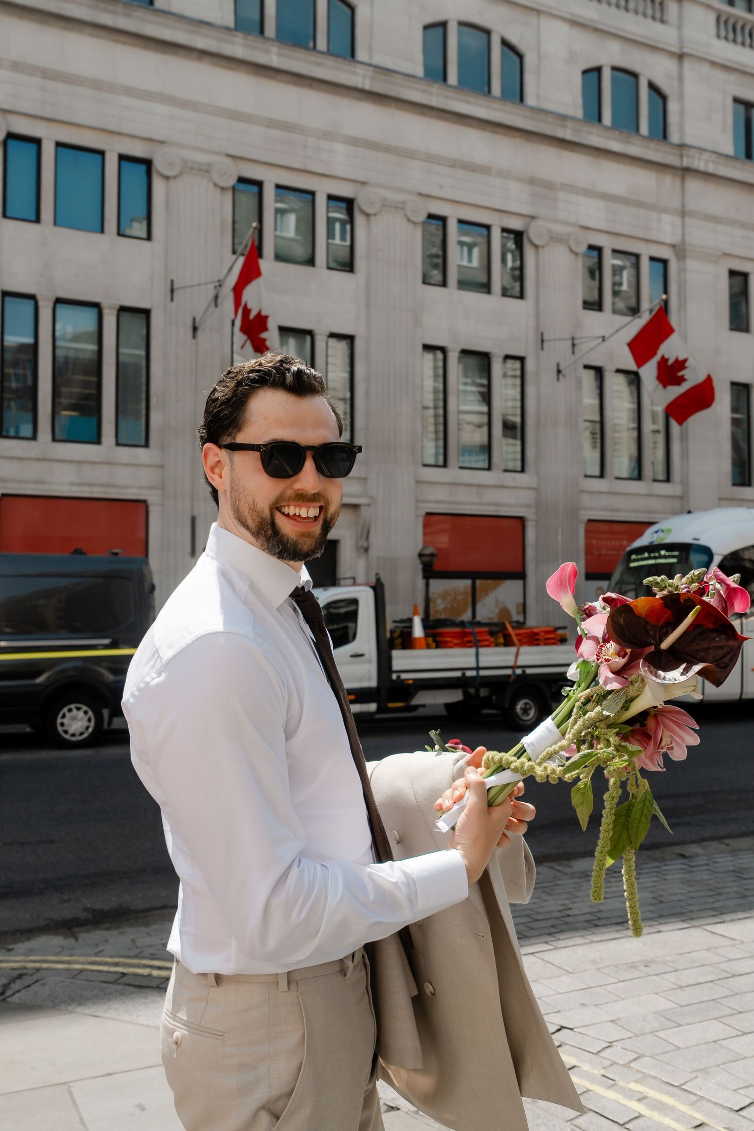 Smiling groom in sunglasses holds bridal bouquet outside a building with Canadian flags in London.