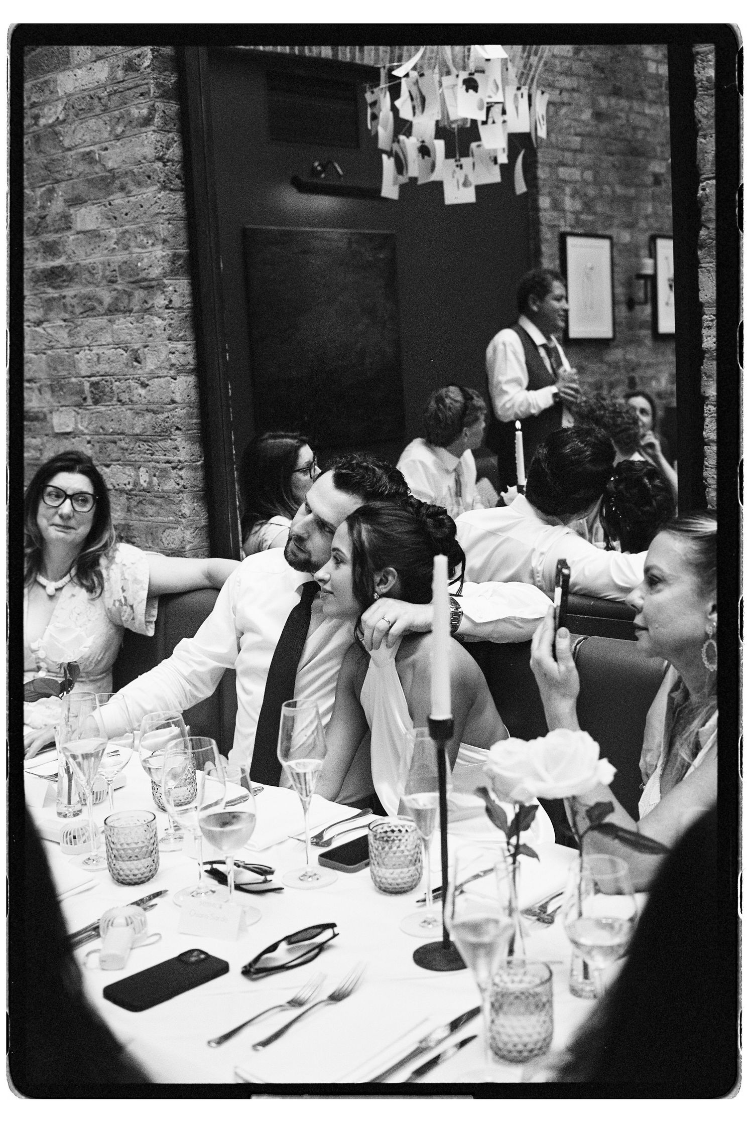 Black and white candid shot of wedding guests laughing and chatting at decorated reception dinner table.