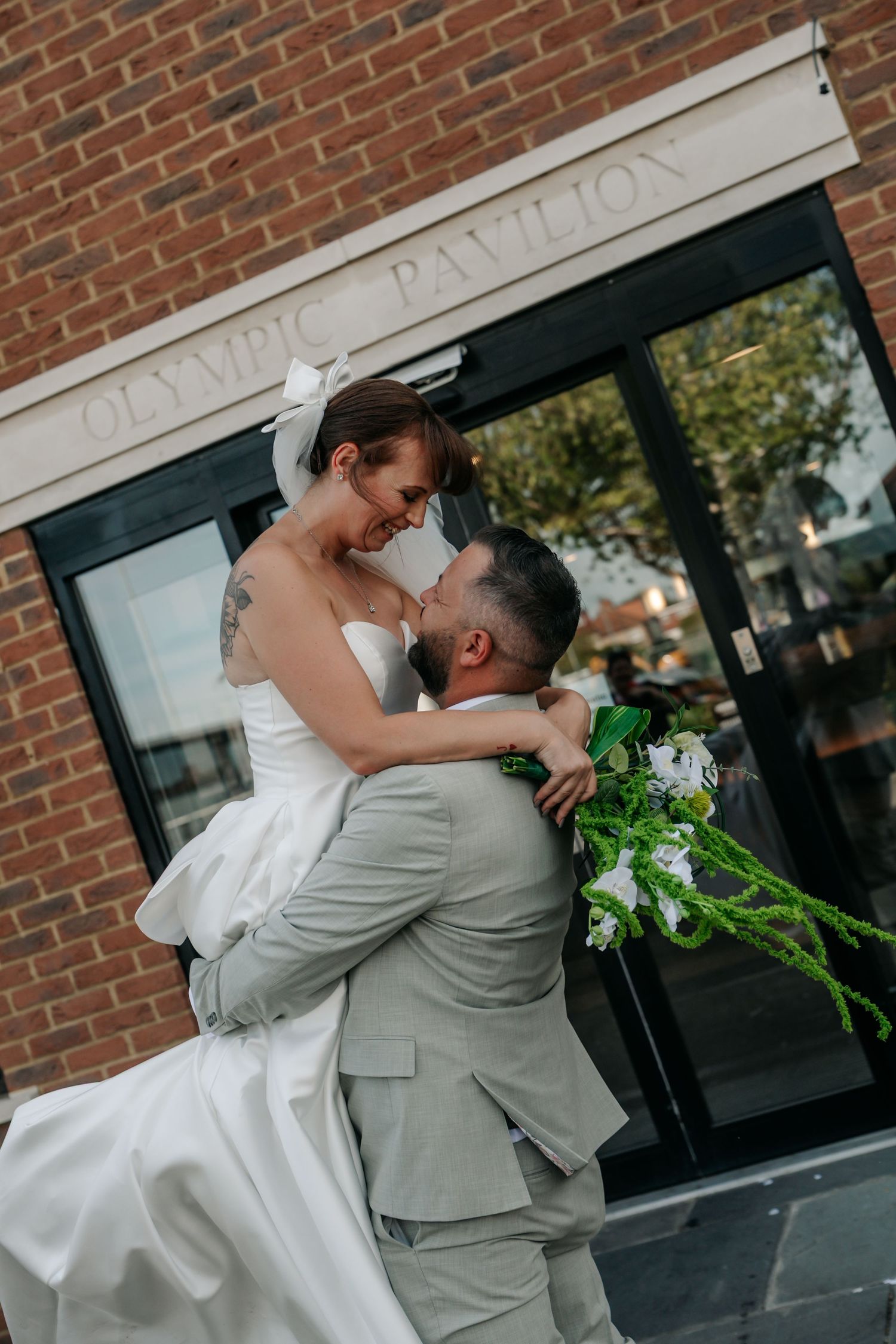 Groom lifts bride in white gown outside Olympic Pavilion, sharing a joyful moment with green floral bouquet.