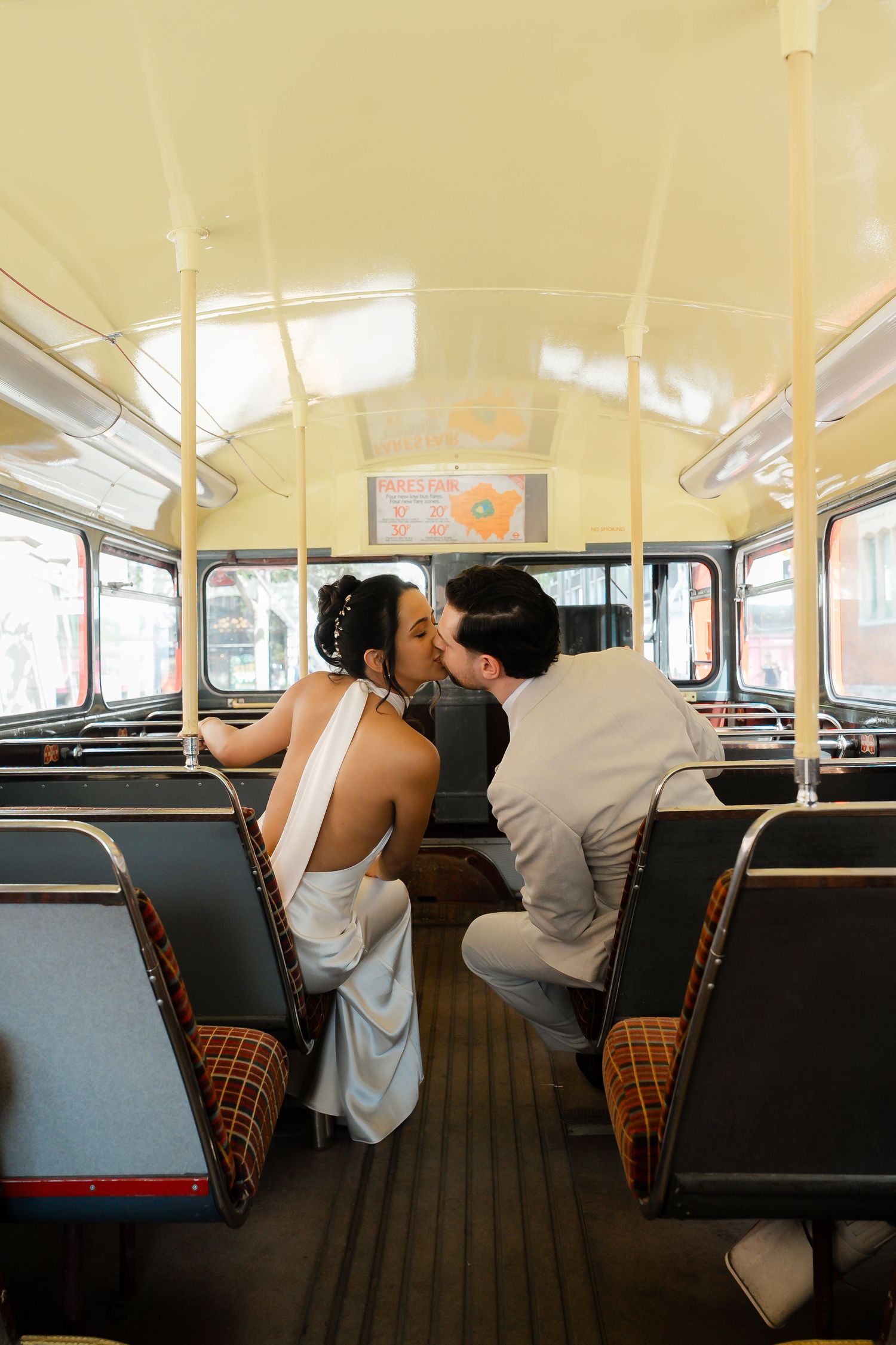 Bride and groom sharing a romantic kiss inside a vintage double-decker bus with retro orange and brown seats.