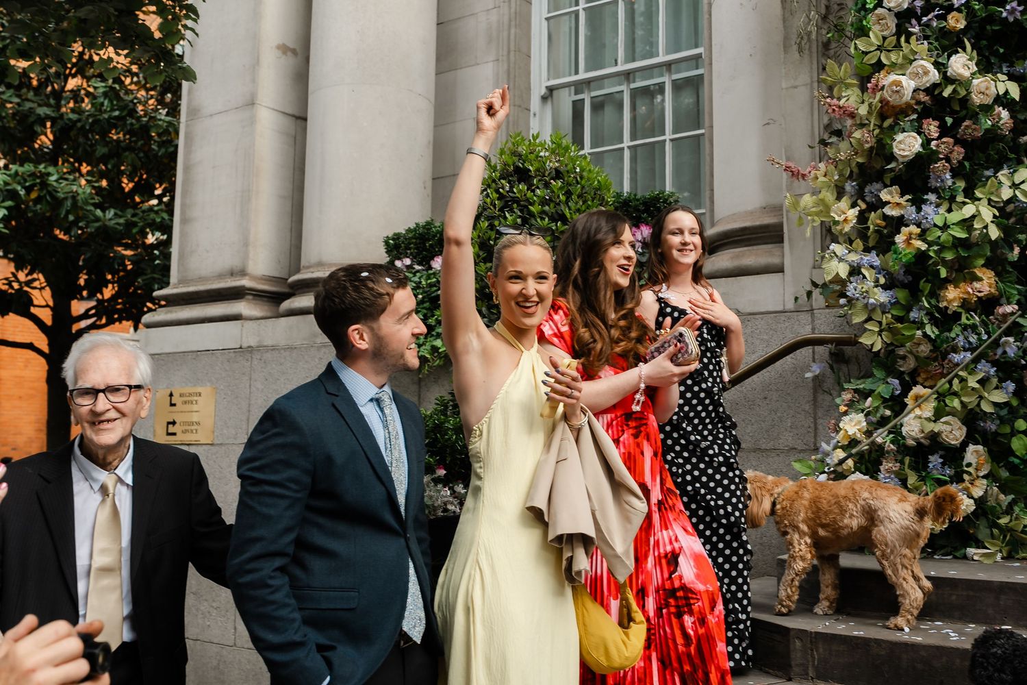 Wedding guests celebrate outside floral-adorned venue as woman raises arm in joy.