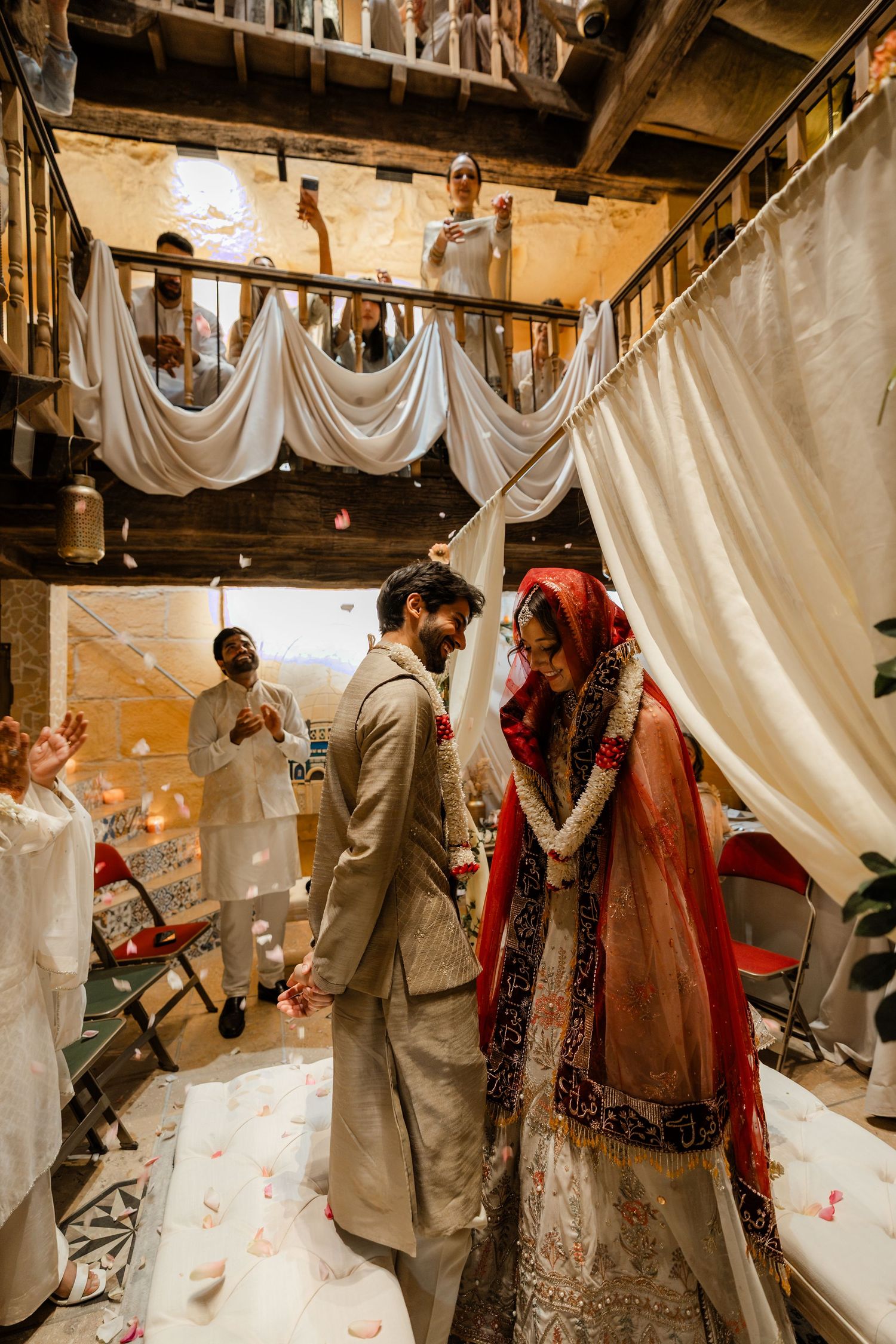 South Asian bride in red dupatta and groom in beige sherwani walk under white drapes at indoor wedding ceremony.