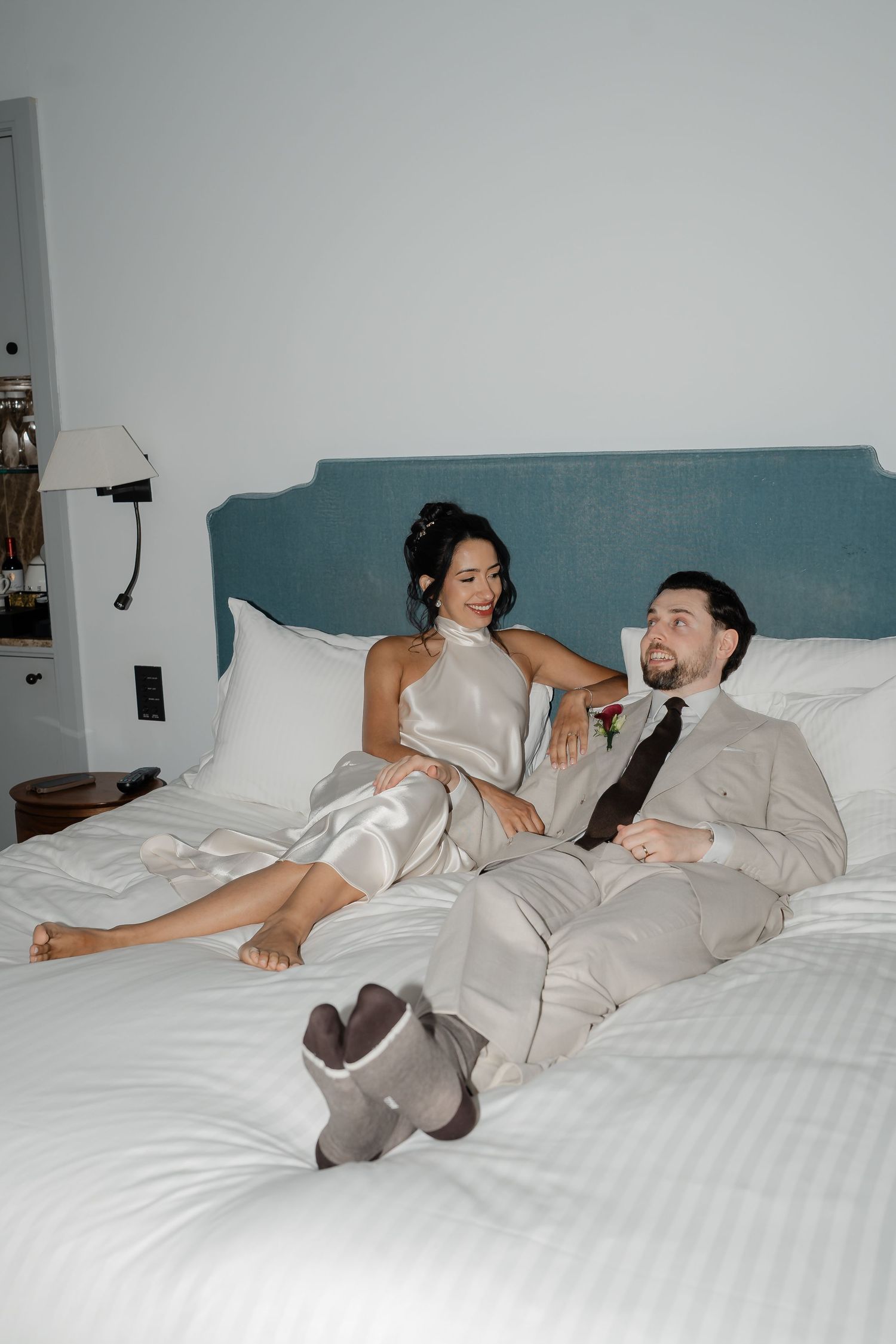 Bride and groom relaxing on a white hotel bed, she in a silk halter dress, he in a beige suit with tie.