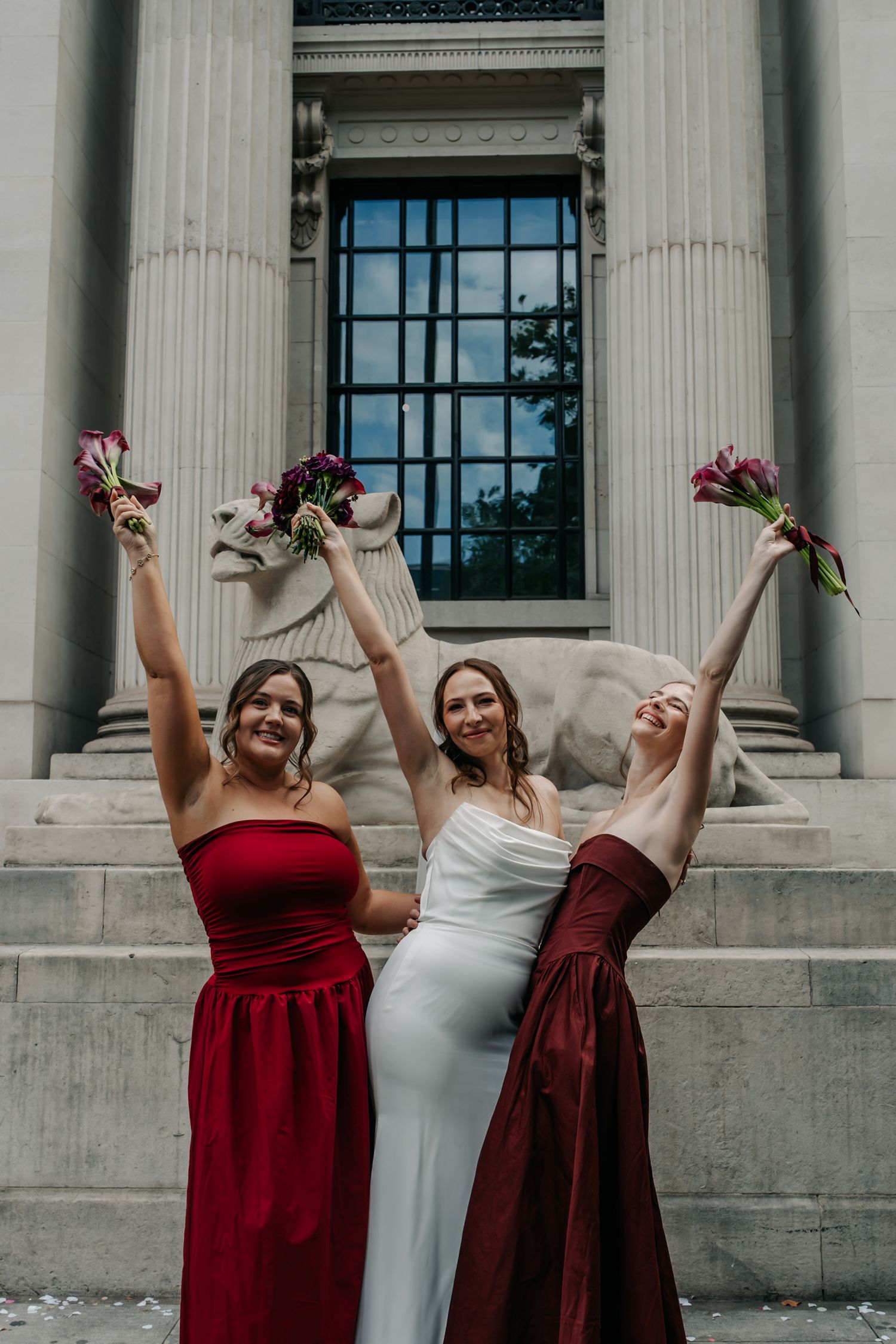 Bride and two bridesmaids in red dresses raise bouquets of purple flowers outside neoclassical building.