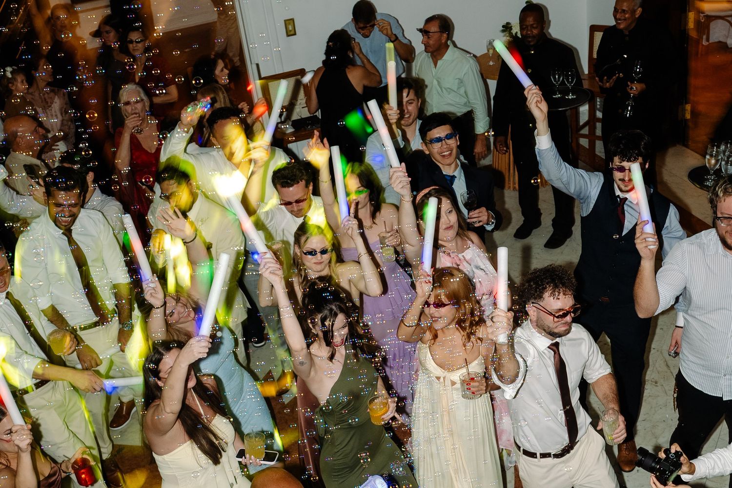 Wedding guests dance with colorful glow sticks during lively reception celebration with confetti.
