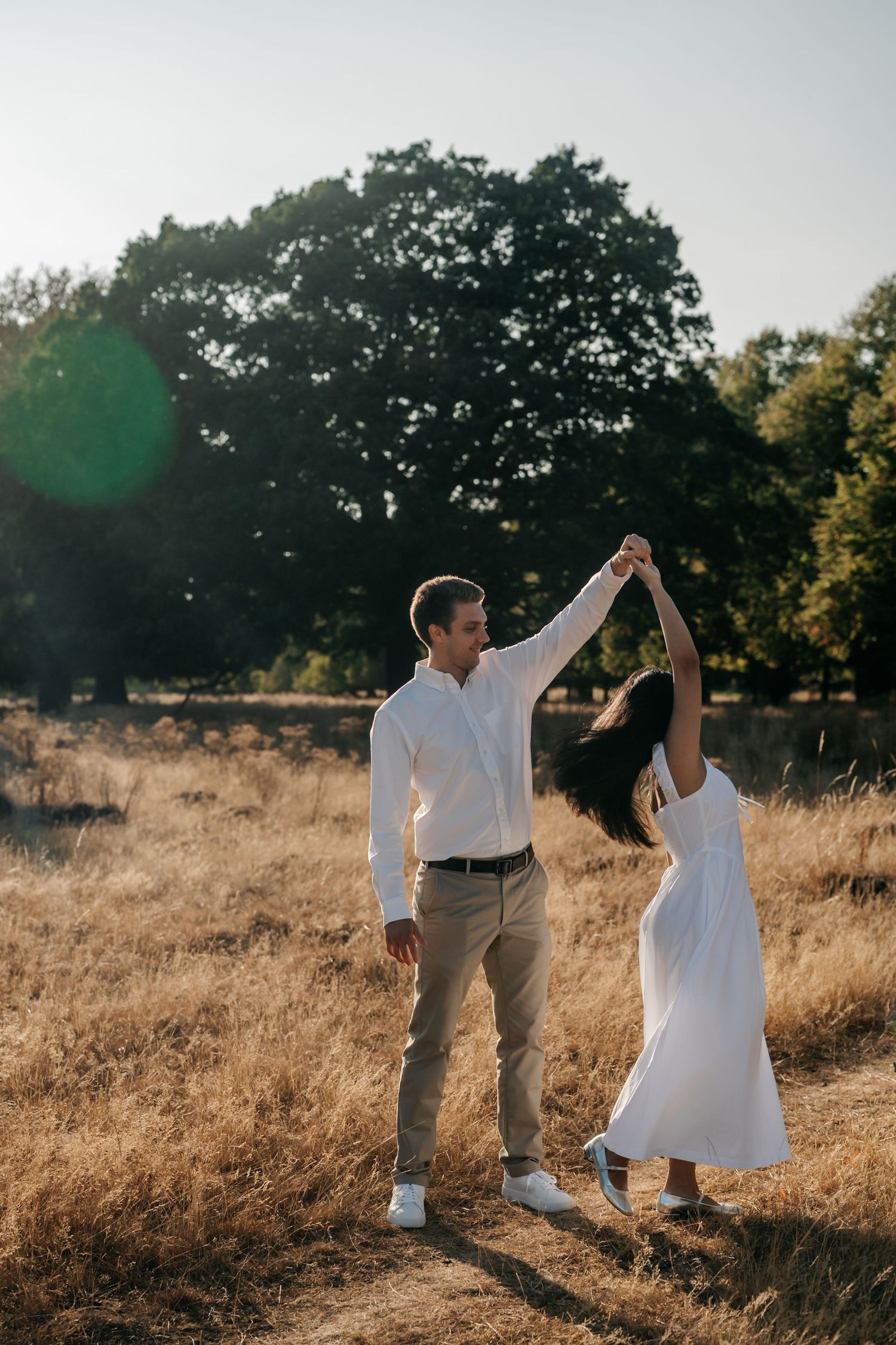 Couple dancing in a golden sunlit field, man in white shirt spinning woman in white dress near large oak tree.