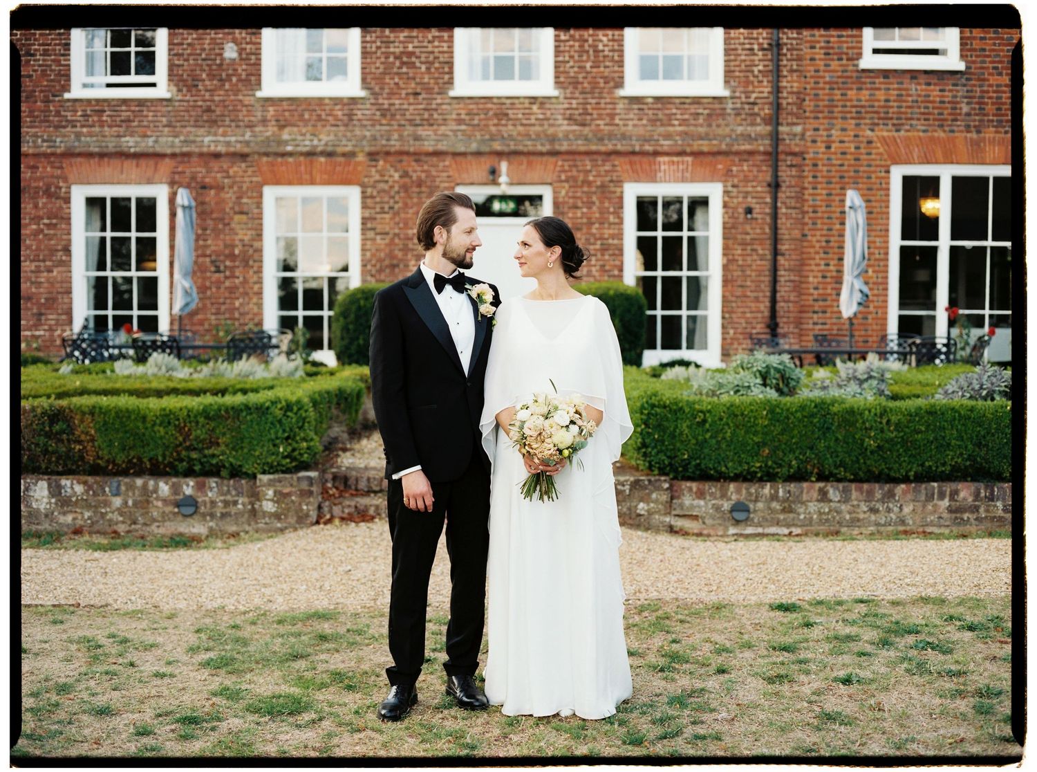 Bride and groom posing in front of a grand brick manor house, bride holding a pastel bouquet in elegant white gown.