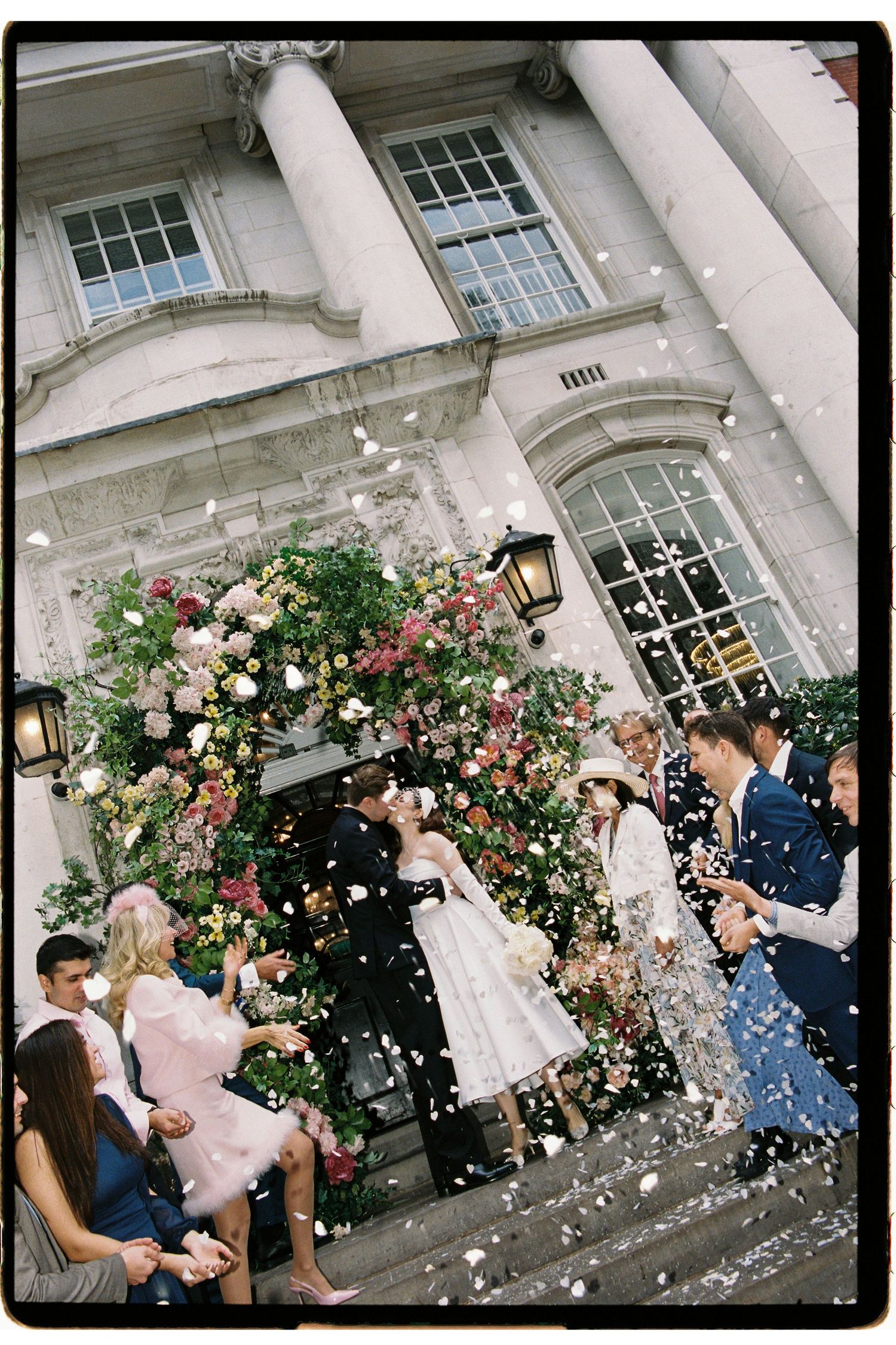 Newlyweds kiss on steps of grand building as guests throw white confetti, surrounded by lush floral arch decorations.