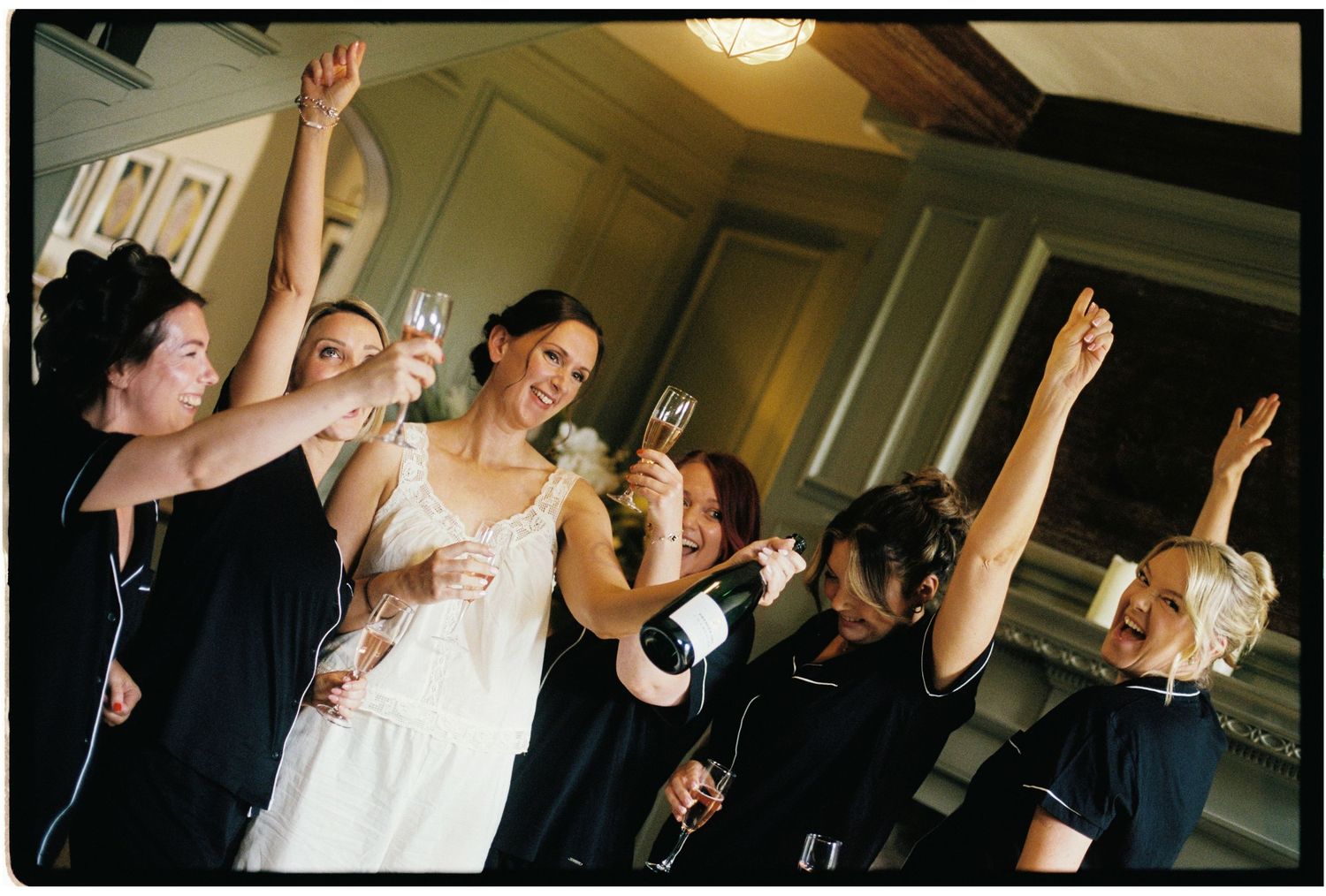 Bride and bridesmaids celebrate with champagne toast, raising glasses and arms joyfully at a wedding reception.