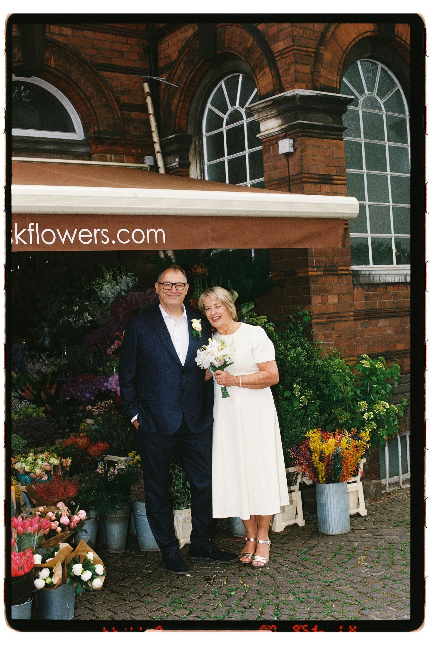 Couple in wedding attire posing outside a flower shop, bride holding bouquet, groom in navy suit, brick building backdrop.