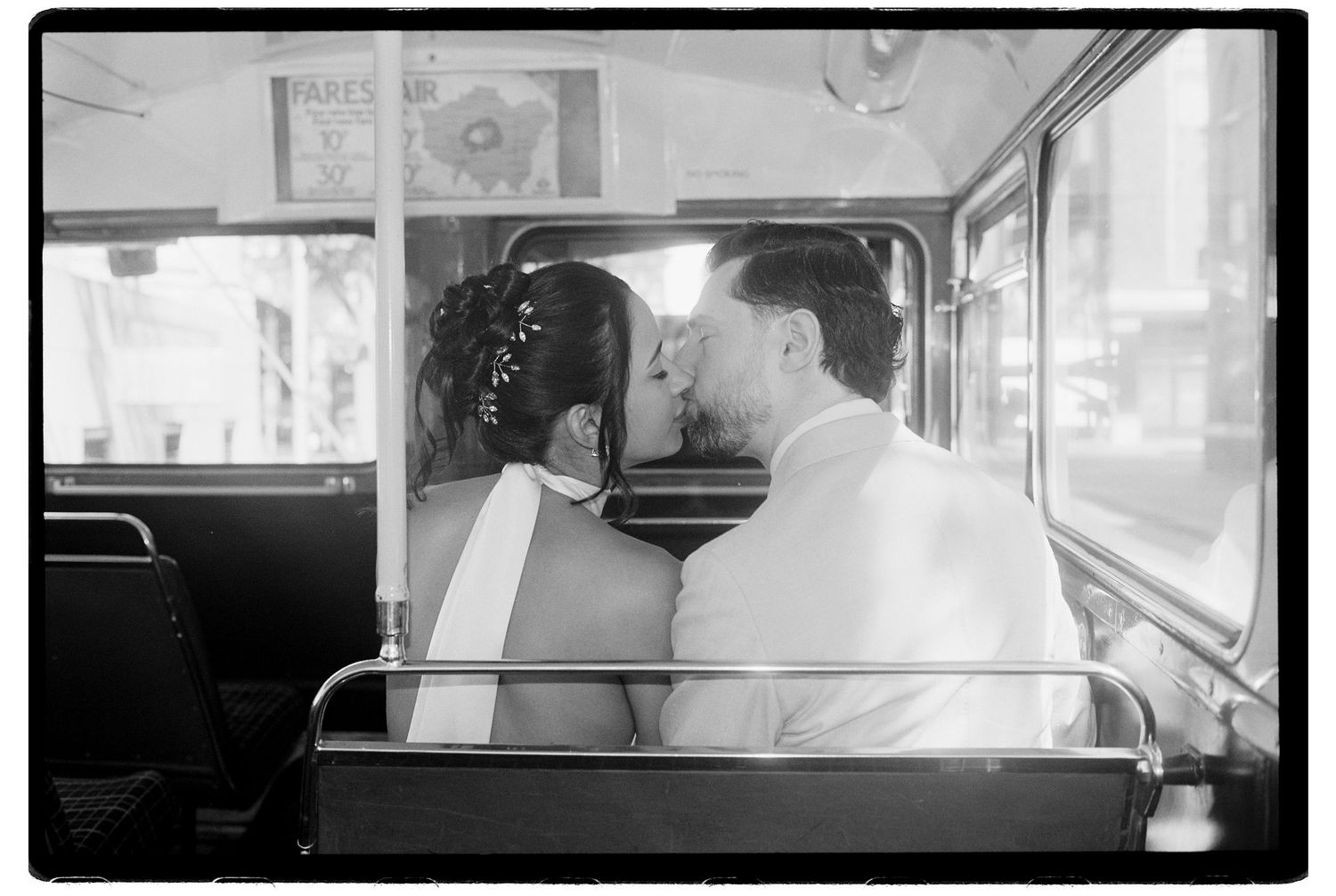 Bride and groom sharing a tender kiss seated on a vintage bus, captured in classic black and white film photography.