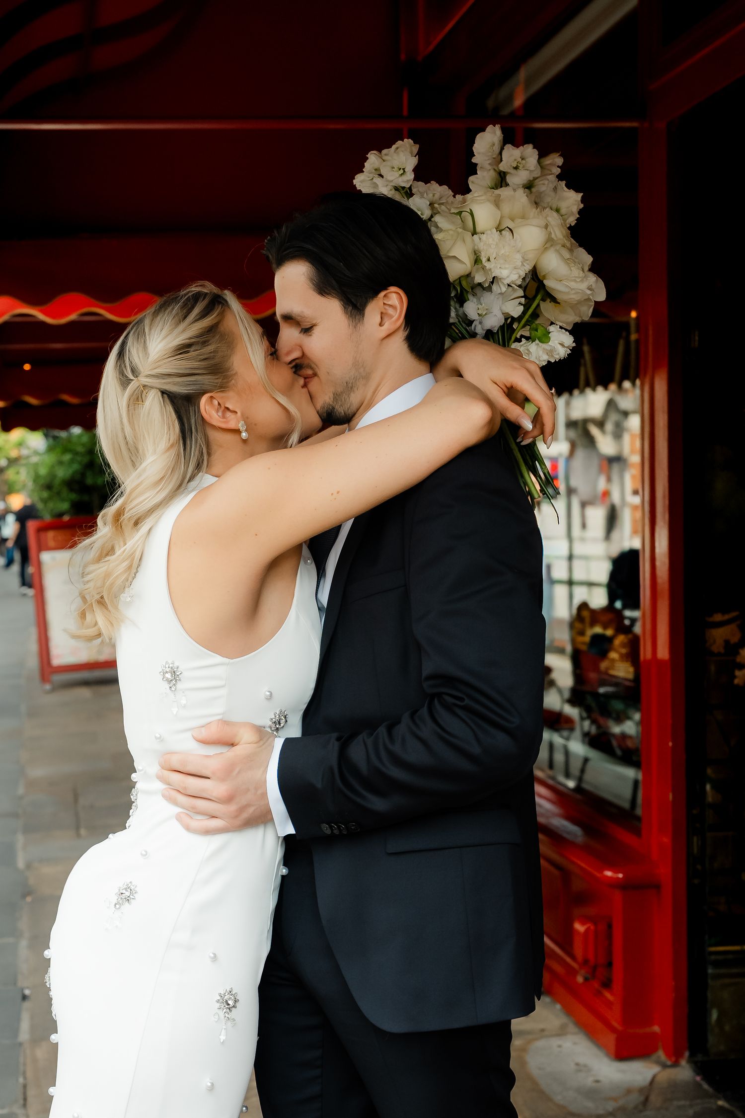Bride and groom share a passionate kiss outside a red-awninged London cafe on their wedding day.
