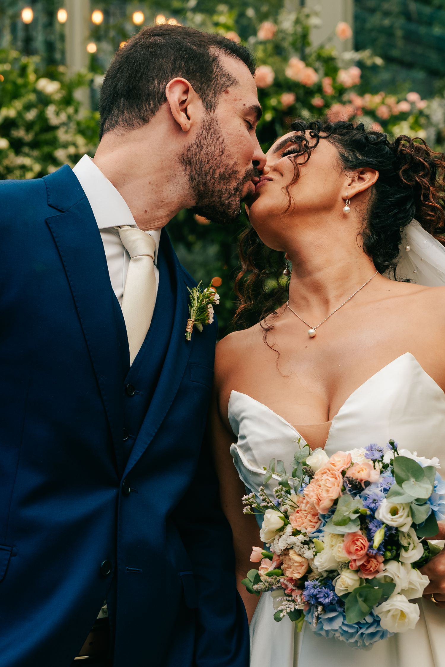Bride and groom share a kiss as bride holds colorful bouquet of peach and blue flowers.