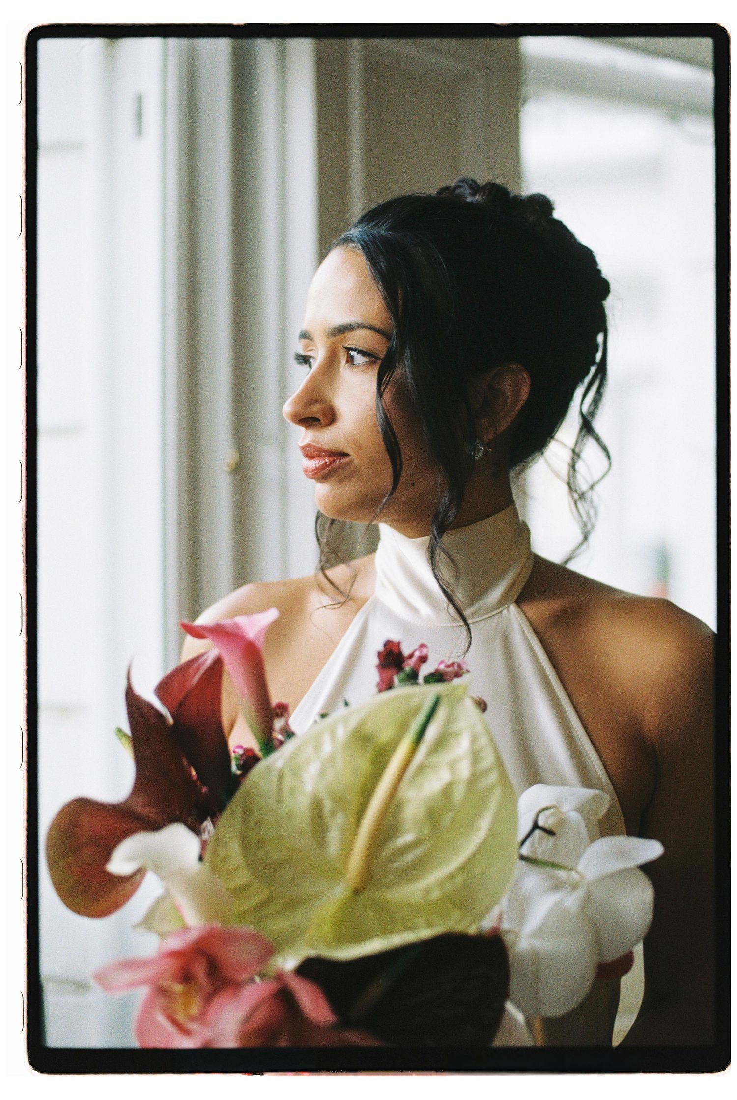 Elegant bride in white halter gown holding tropical floral bouquet gazing out window.