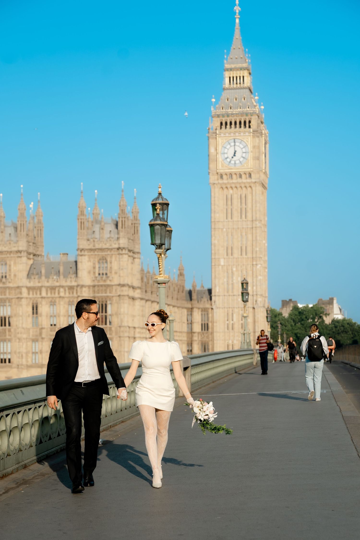 Bride in white mini dress and groom walking hand-in-hand on Westminster Bridge near Big Ben.