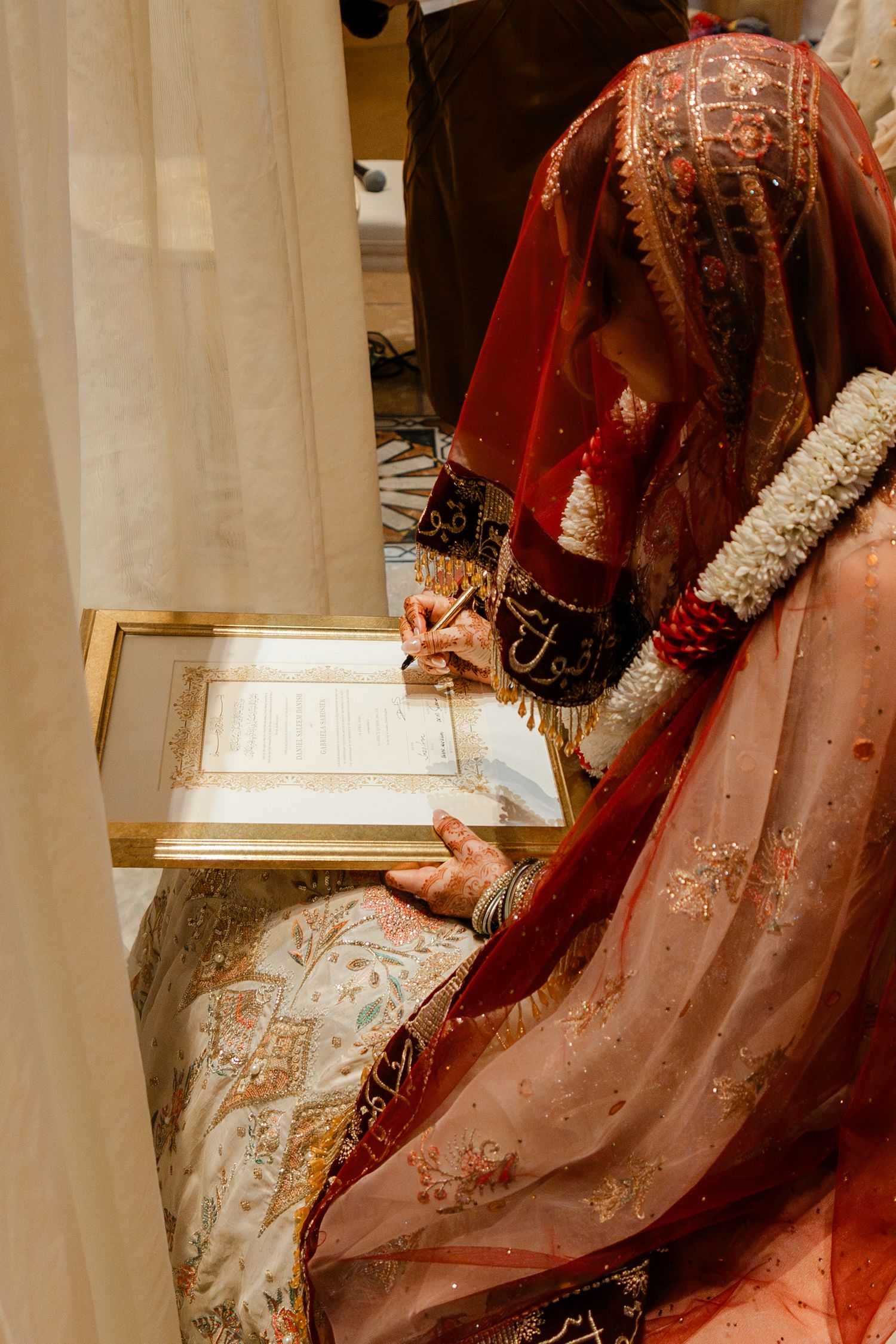 Bride in red embroidered bridal attire signing wedding document framed in gold during traditional ceremony.
