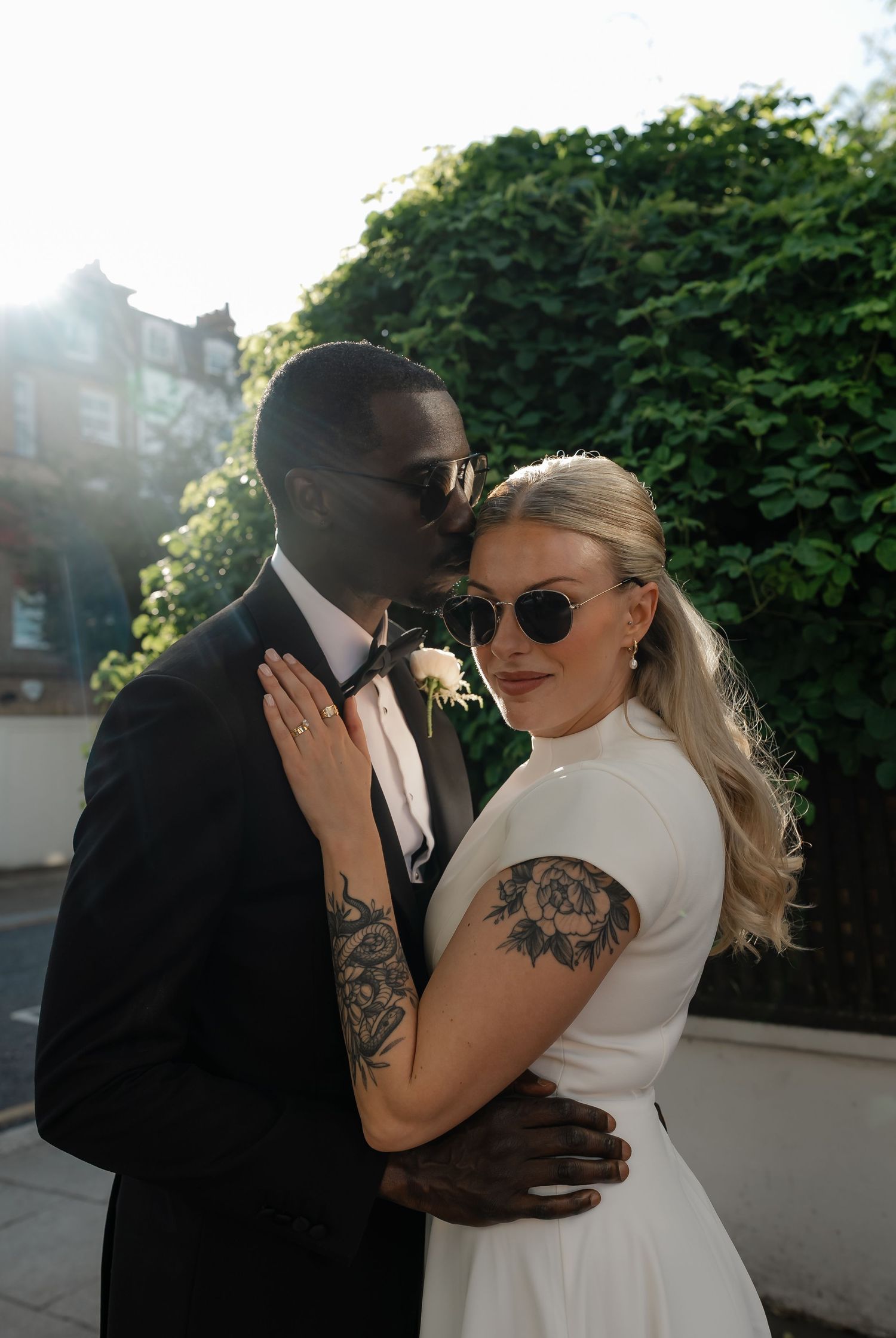 Interracial newlywed couple sharing a tender moment outdoors with sunglasses in golden sunlight.