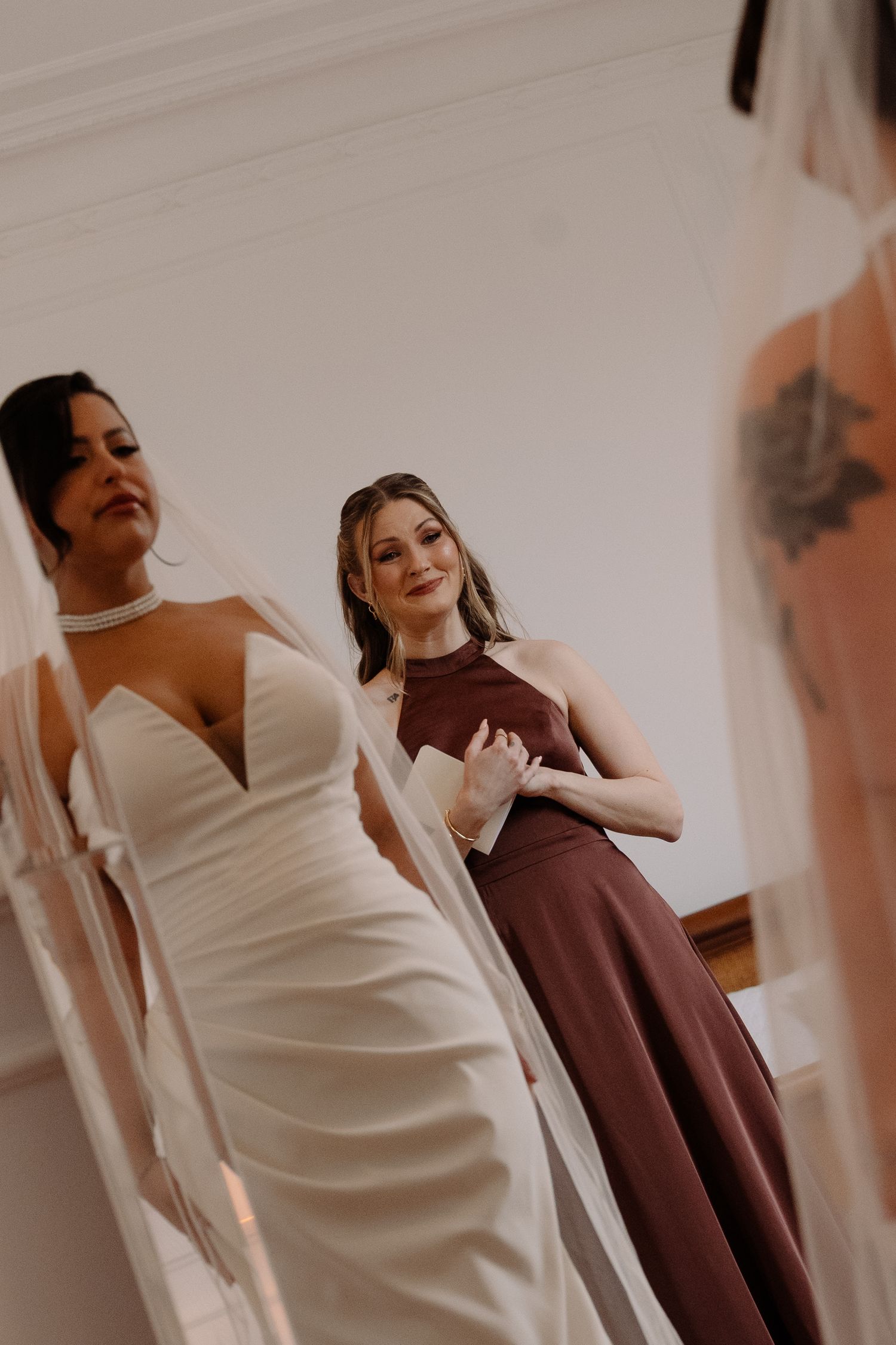 Bridesmaid in brown dress admires bride in white halter gown during wedding preparations.