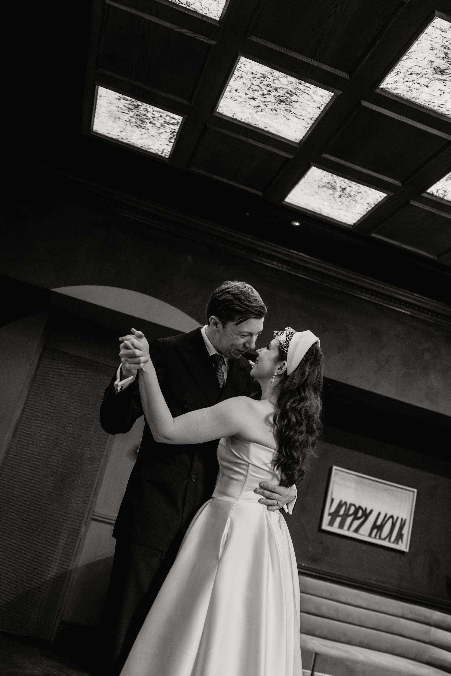 Black and white photo of a bride and groom sharing their first dance, kissing beneath a skylight ceiling.