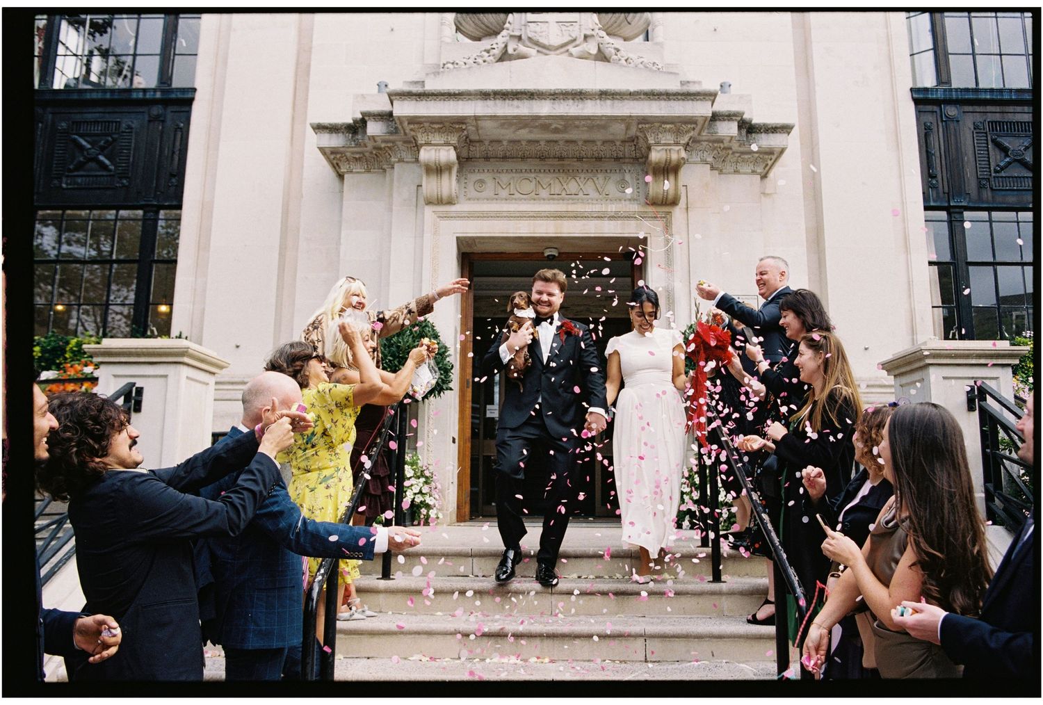 Newlywed couple exits a grand building as guests shower them with colorful confetti at a joyful wedding celebration.