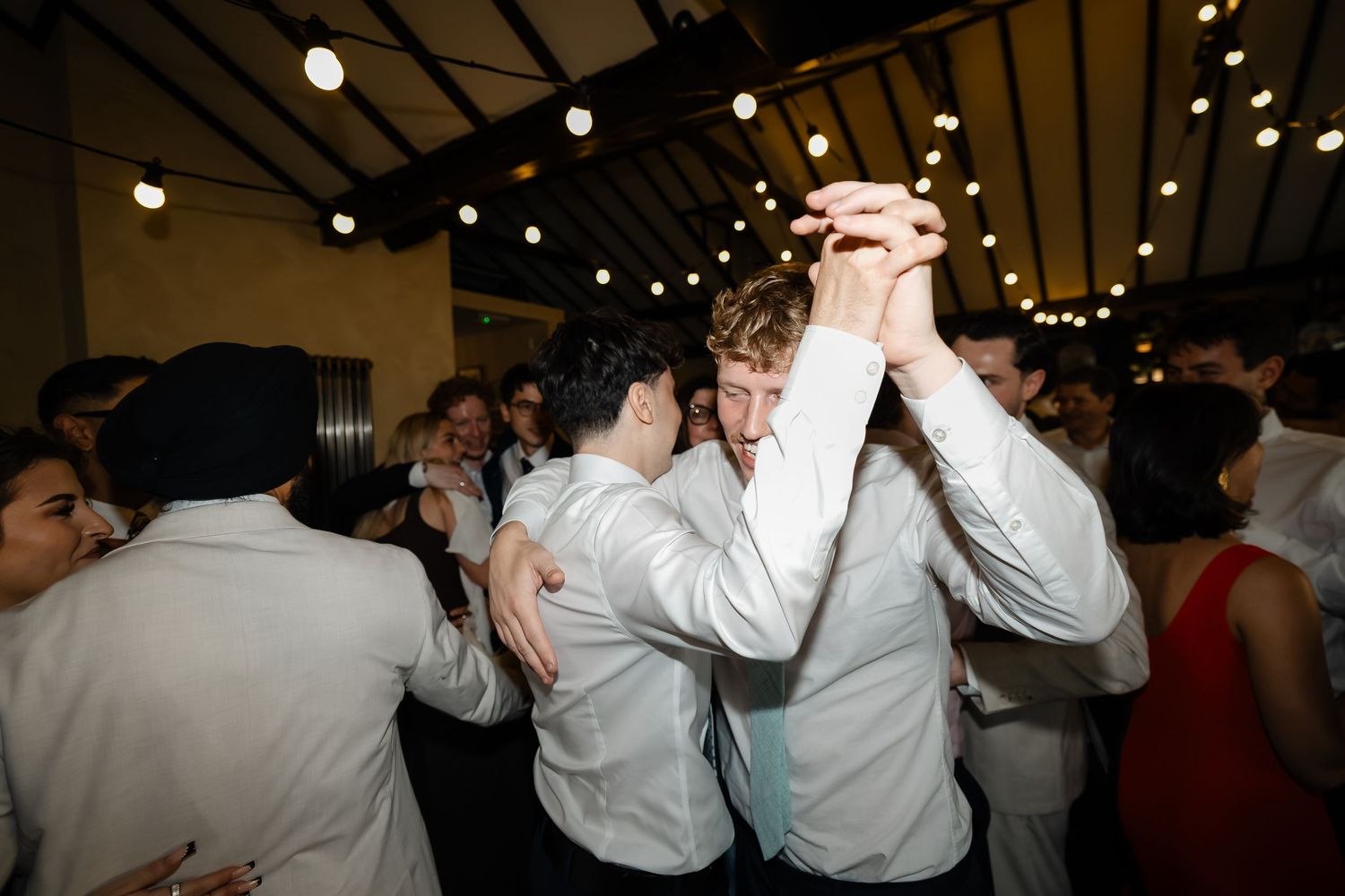 Wedding guests dance energetically together under string lights at a lively reception party.