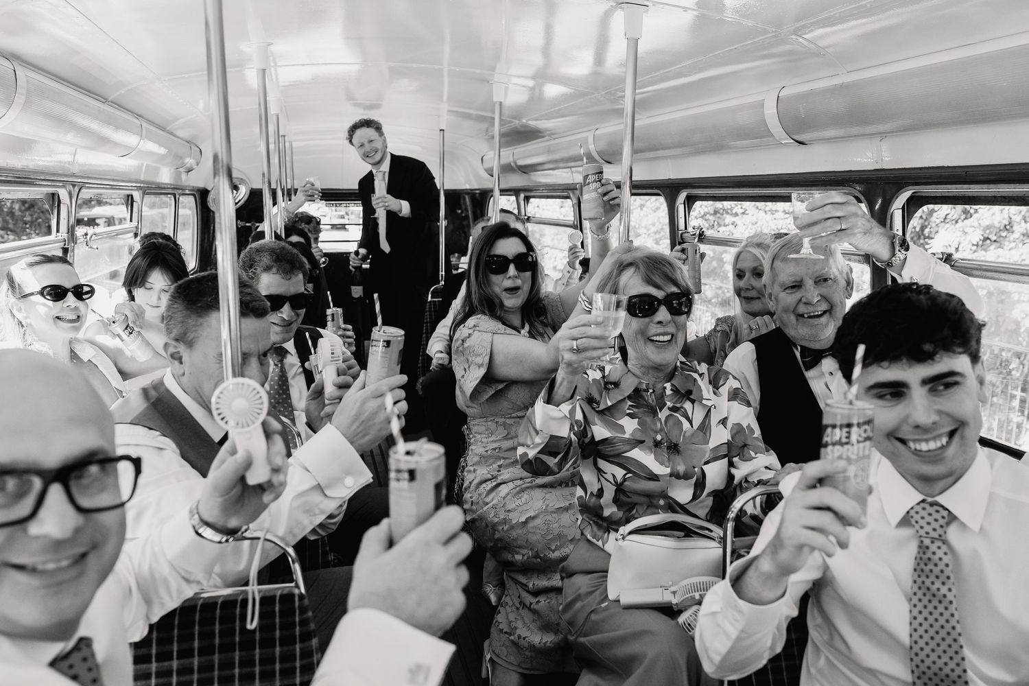Black and white photo of cheerful wedding guests raising glasses and celebrating inside a vintage double-decker bus.