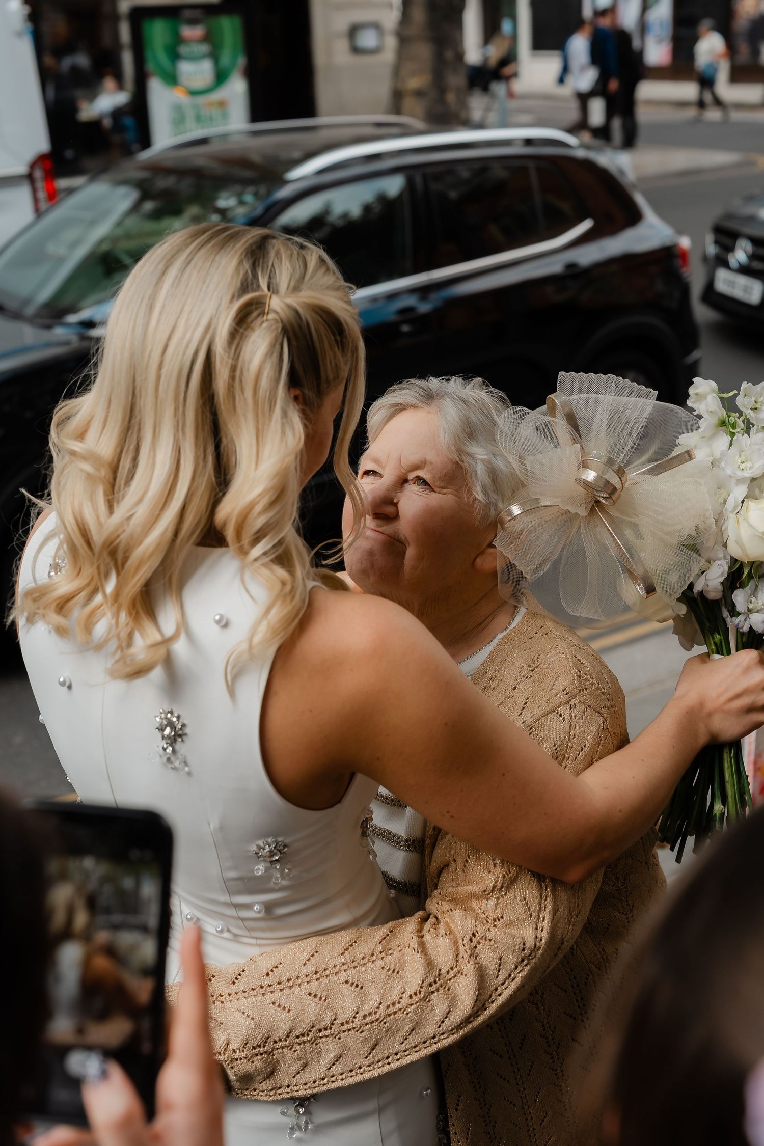 Bride in white gown embraces elderly woman holding white floral bouquet on street.