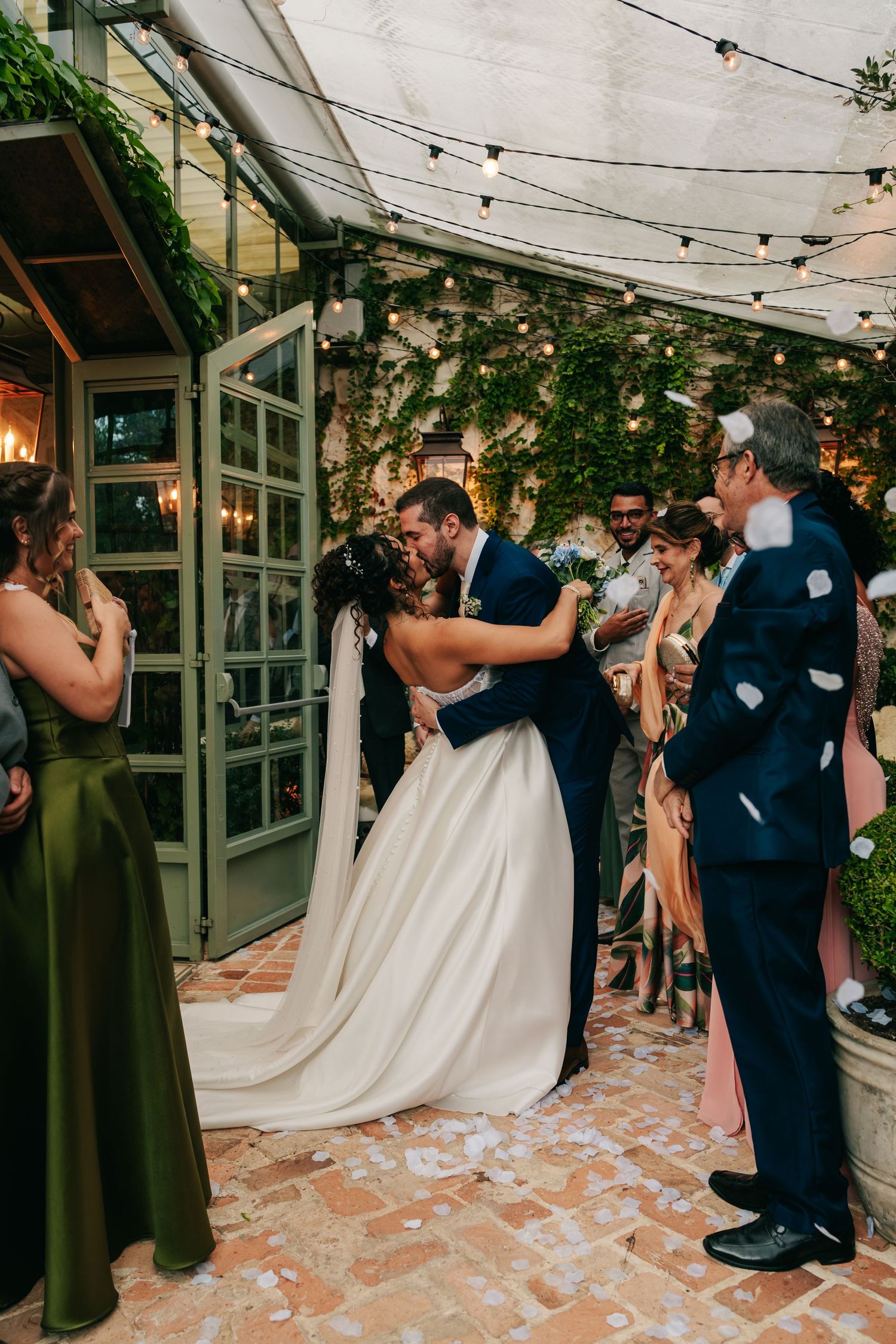 Bride and groom share romantic kiss during petal toss at ivy-covered outdoor wedding venue.