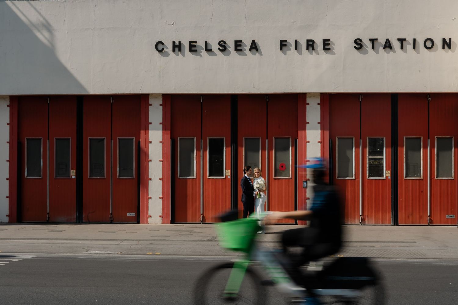 Bride and groom pose in front of Chelsea Fire Station red doors as a cyclist passes by.
