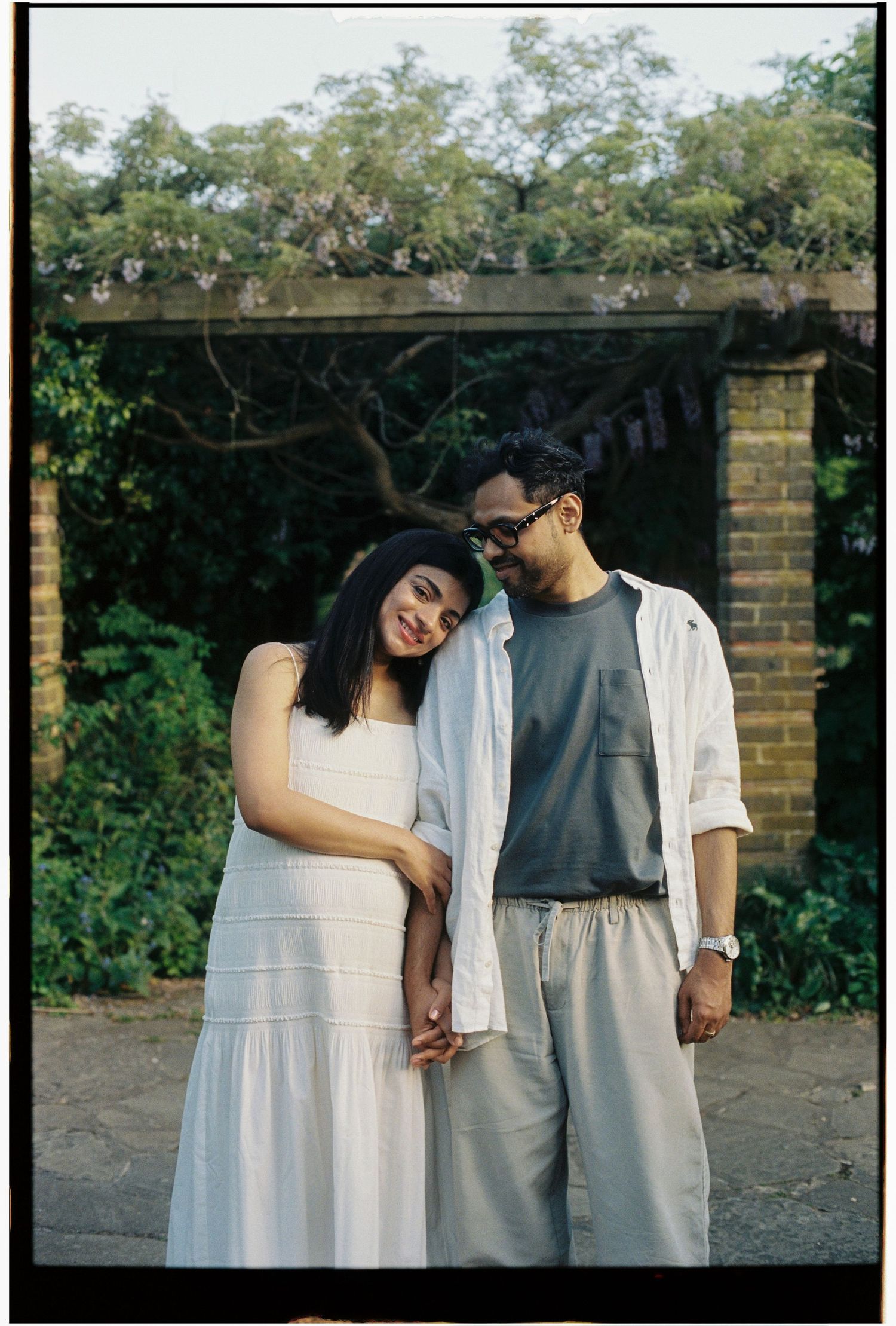 Pregnant couple embracing near brick garden wall, woman in white dress leaning on man's shoulder.