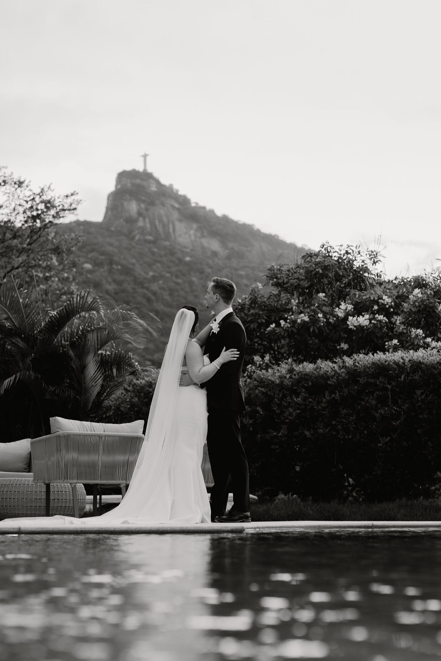 Bride and groom embrace poolside with Christ the Redeemer statue on mountain in black and white.