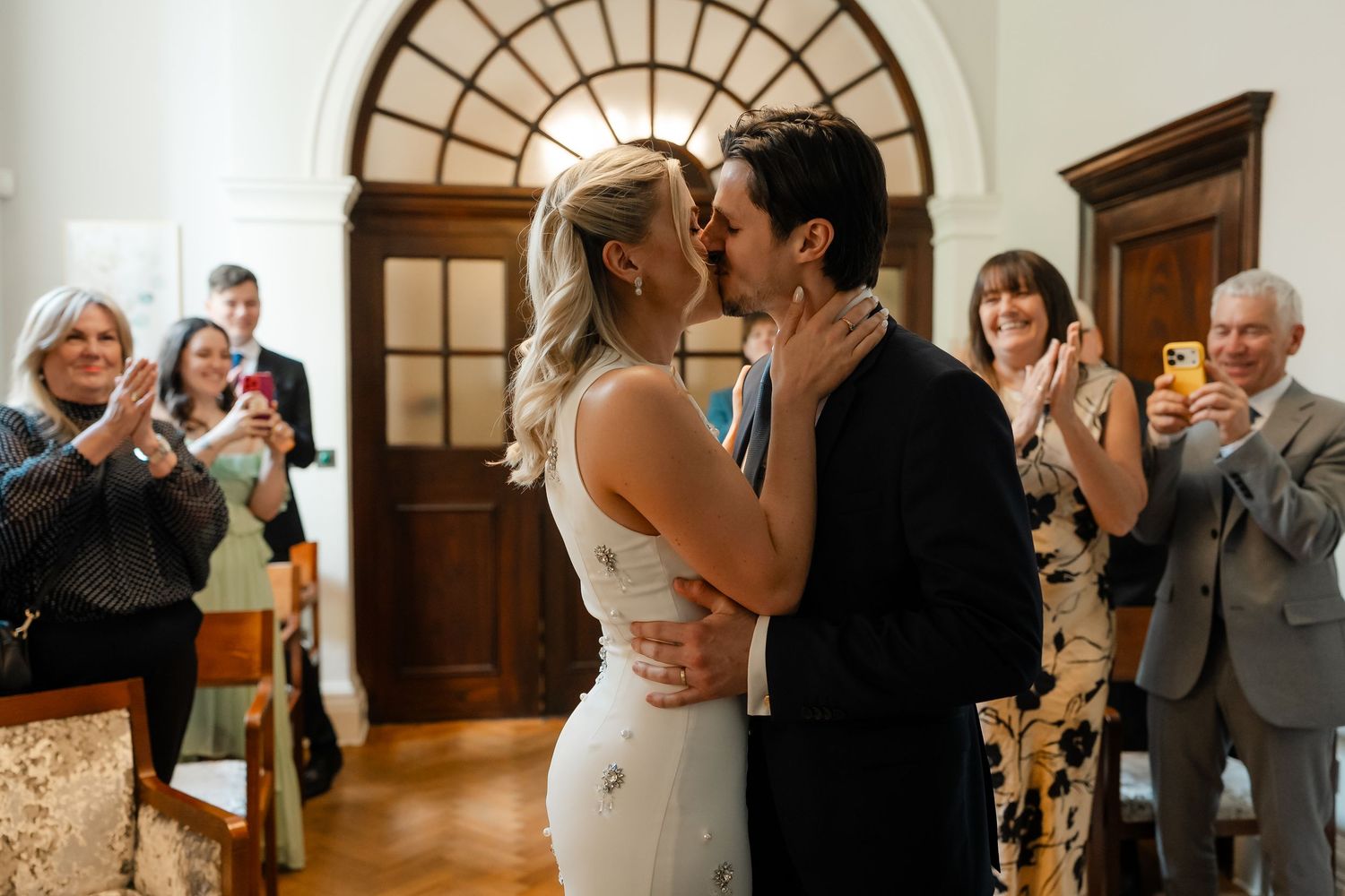 Newlyweds share their first kiss as married couple while guests applaud and photograph the moment.