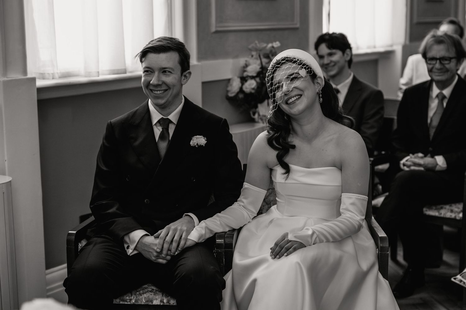 Black and white photo of a smiling bride and groom seated together during their wedding ceremony.