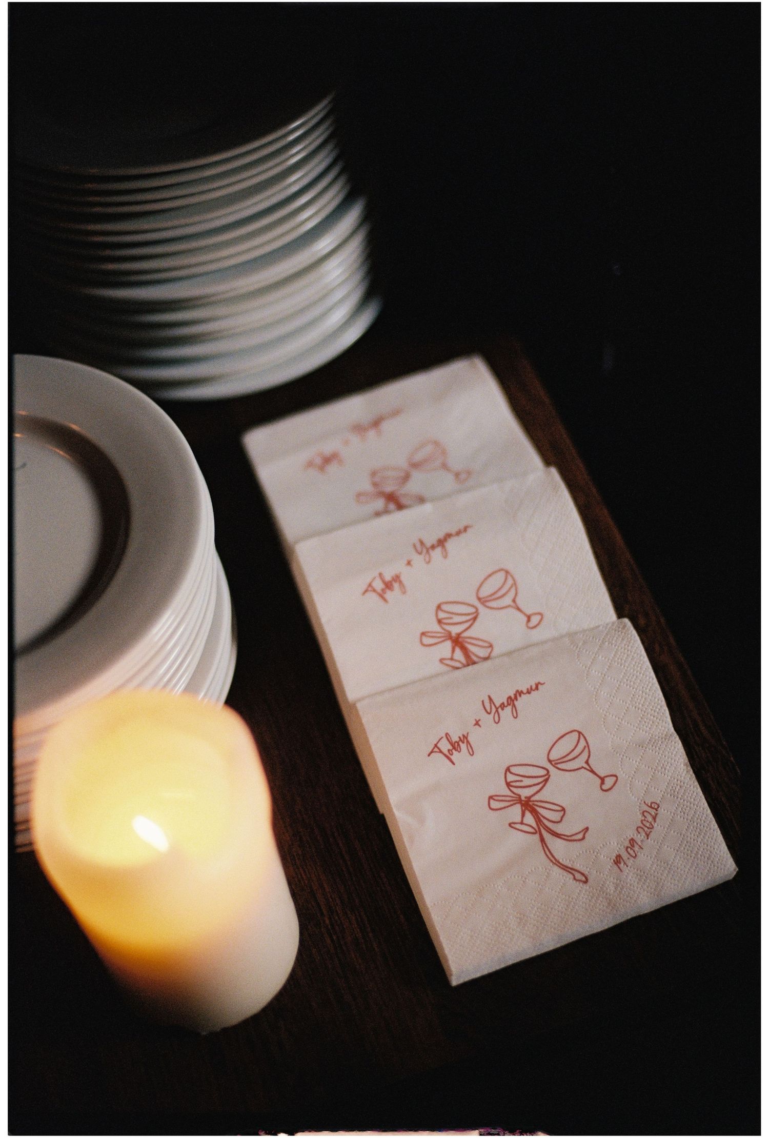 Personalized wedding cocktail napkins with red monogram and wine glass design displayed on wooden tray beside candle and stacked plates.