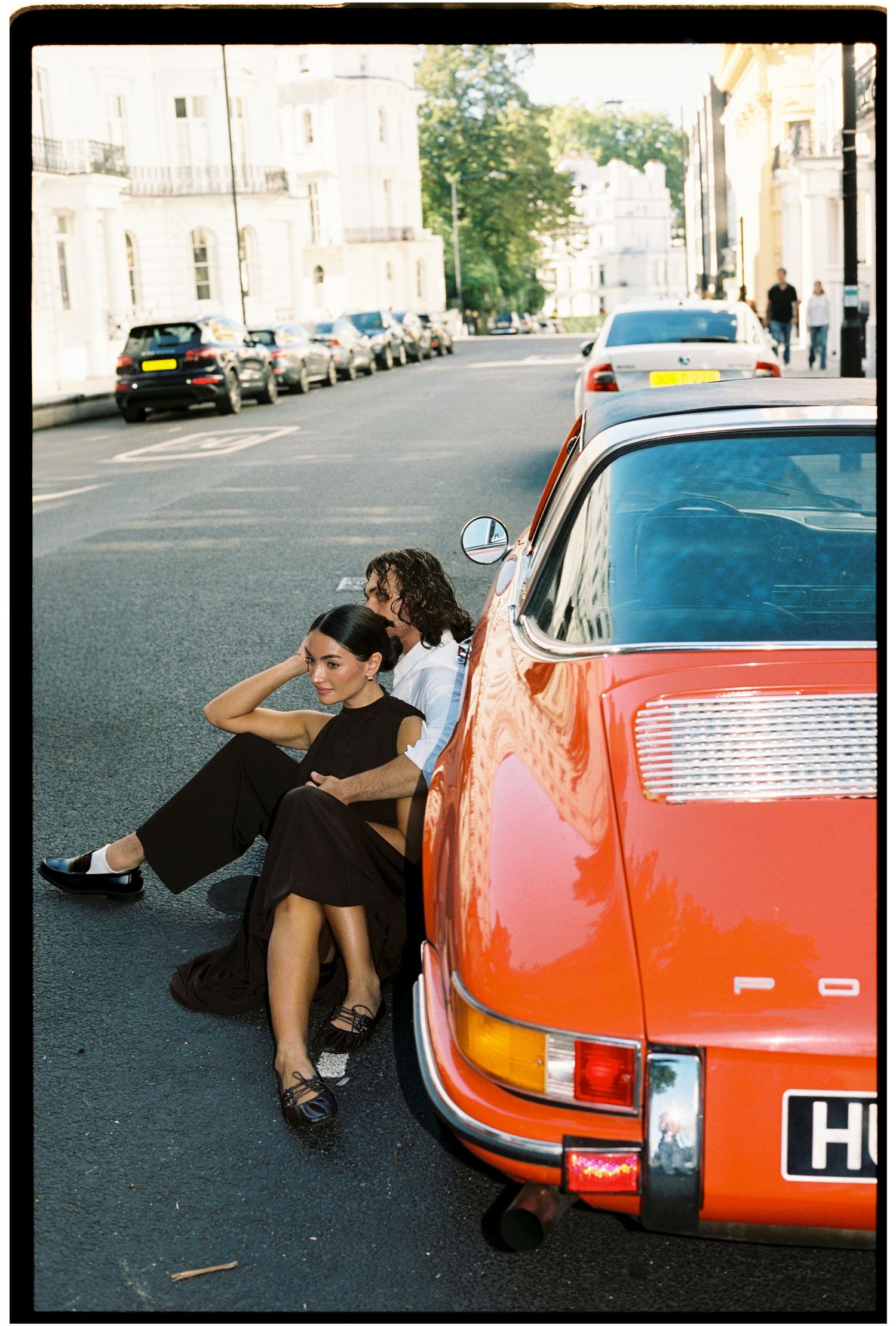 Two people sitting against a vintage orange Porsche on a London street, woman in black dress with loafers, film photography aesthetic.