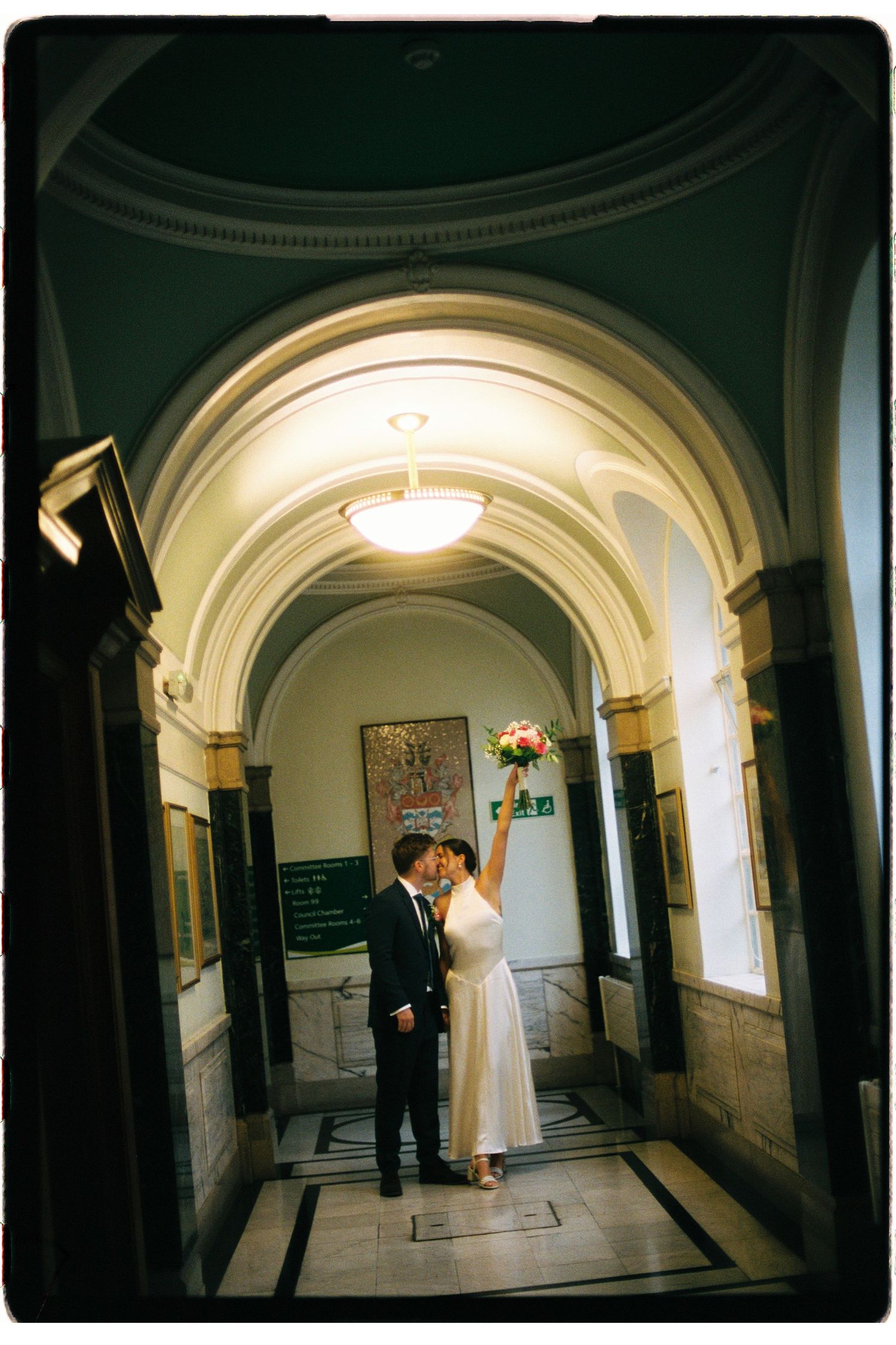 Bride and groom kissing under elegant arched hallway, bride holding bouquet aloft in grand historic building.