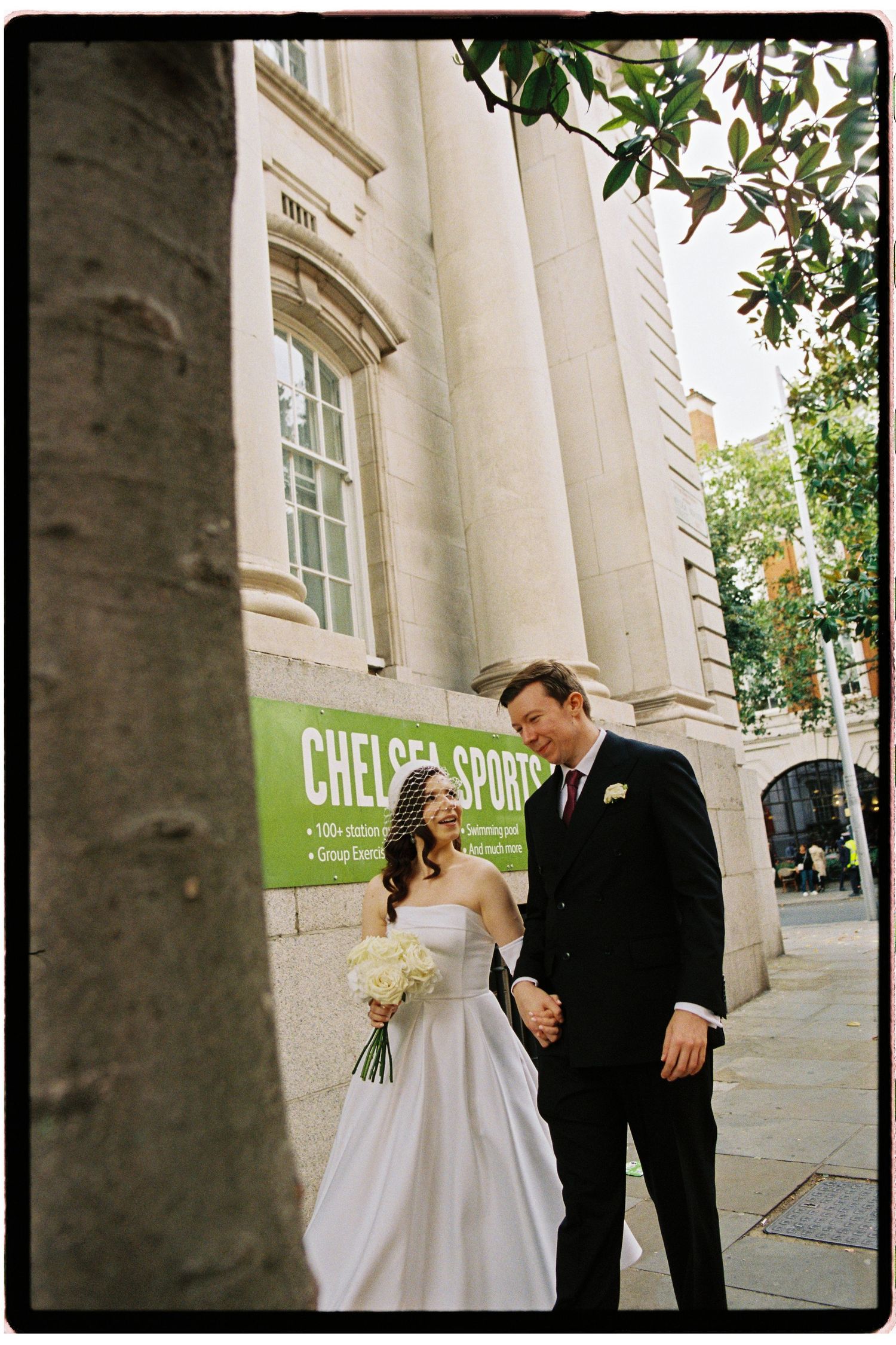 Bride and groom holding hands outside Chelsea Sports building, bride in white strapless gown holding bouquet.